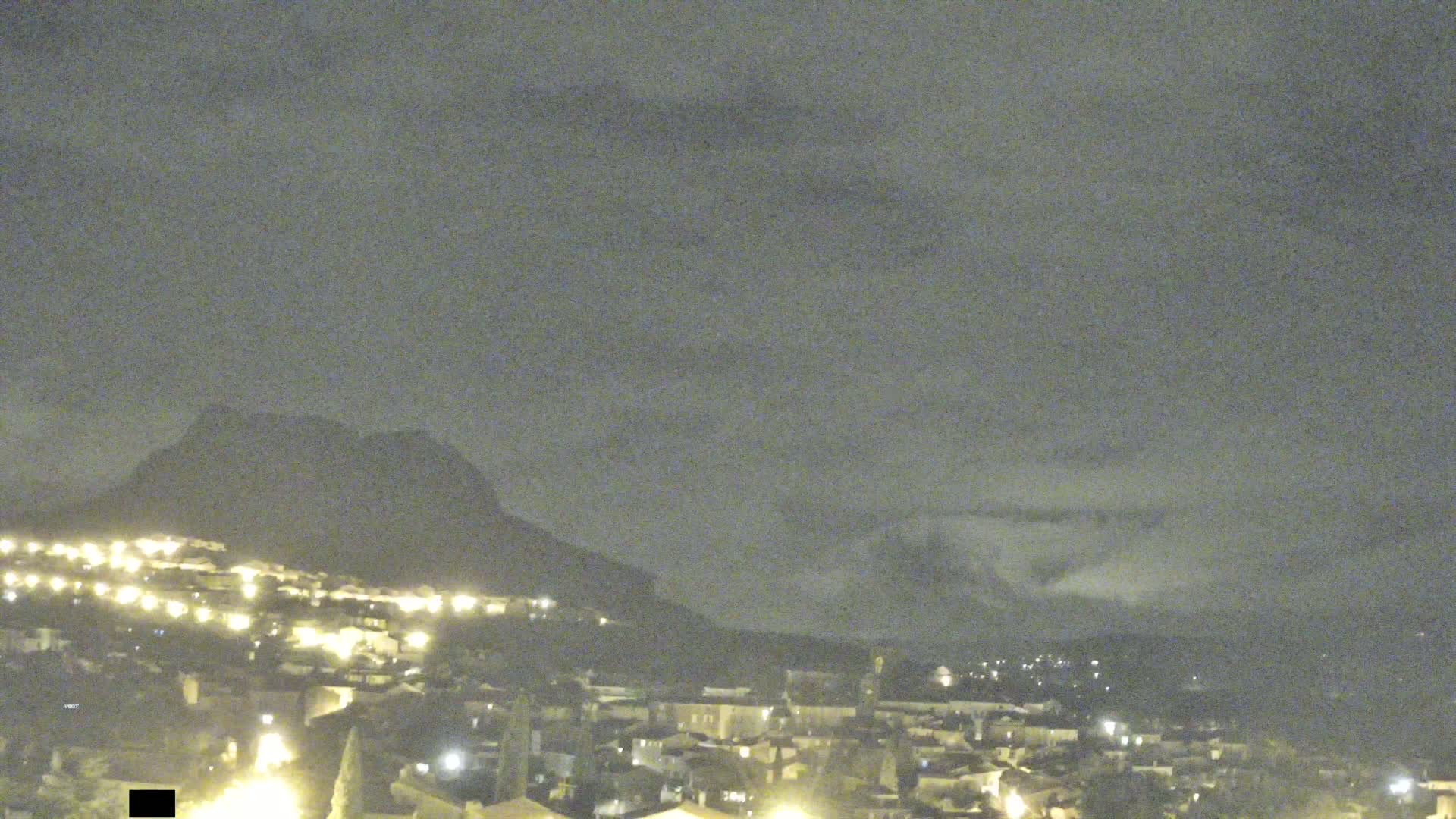 A nighttime view of a town nestled at the base of a mountain under a cloudy sky.