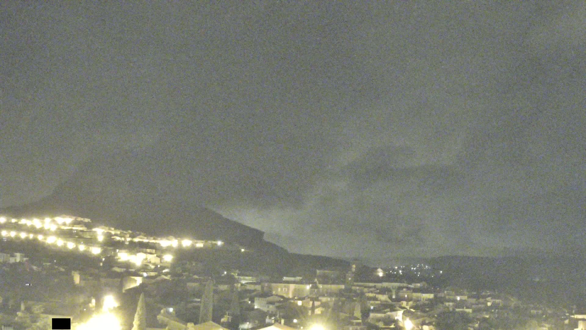 A nighttime view of a town nestled at the base of a hill under a cloudy sky.