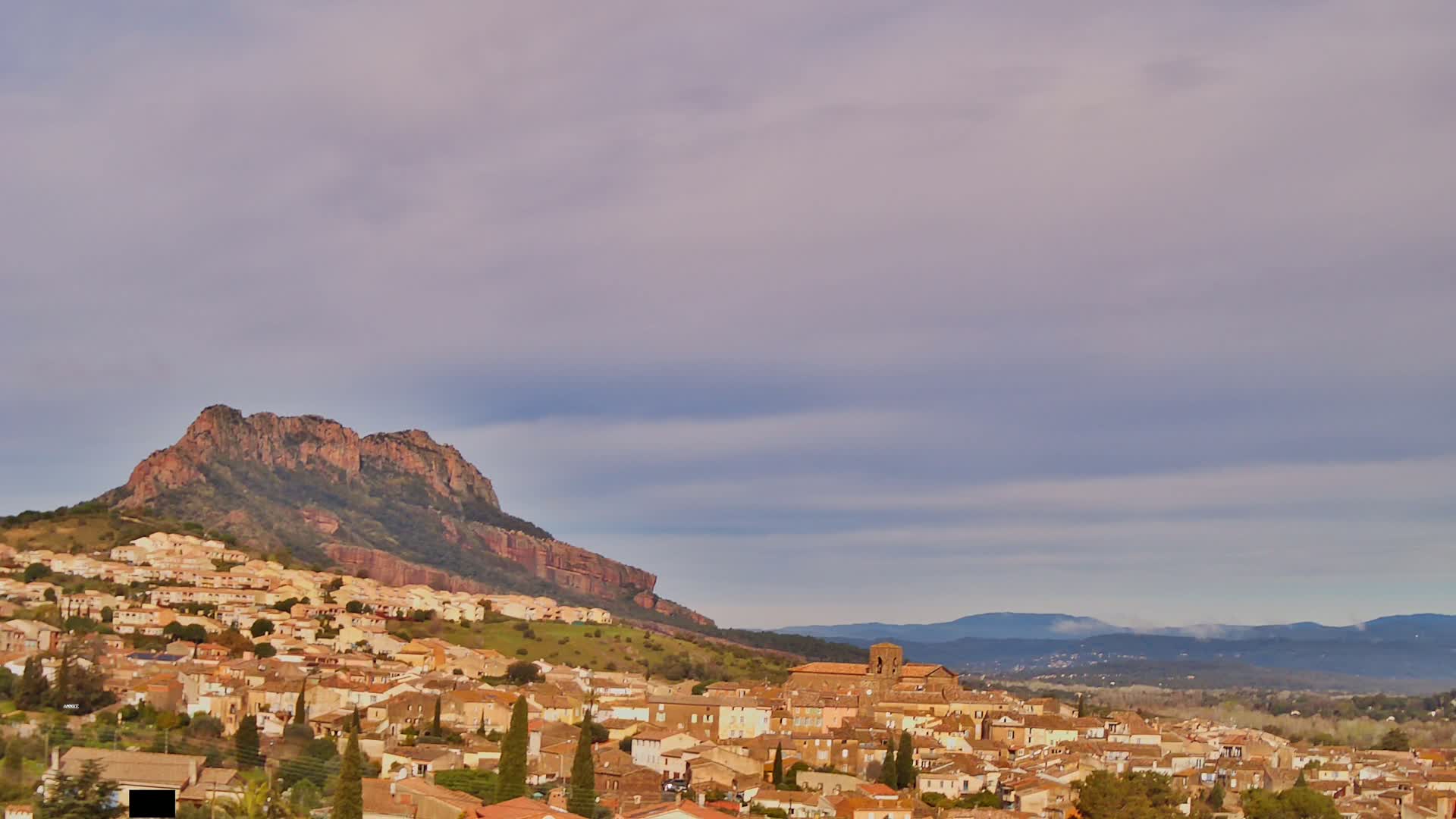 A sprawling town of tan buildings nestled against a large, reddish-brown hill under a pale, cloudy sky.