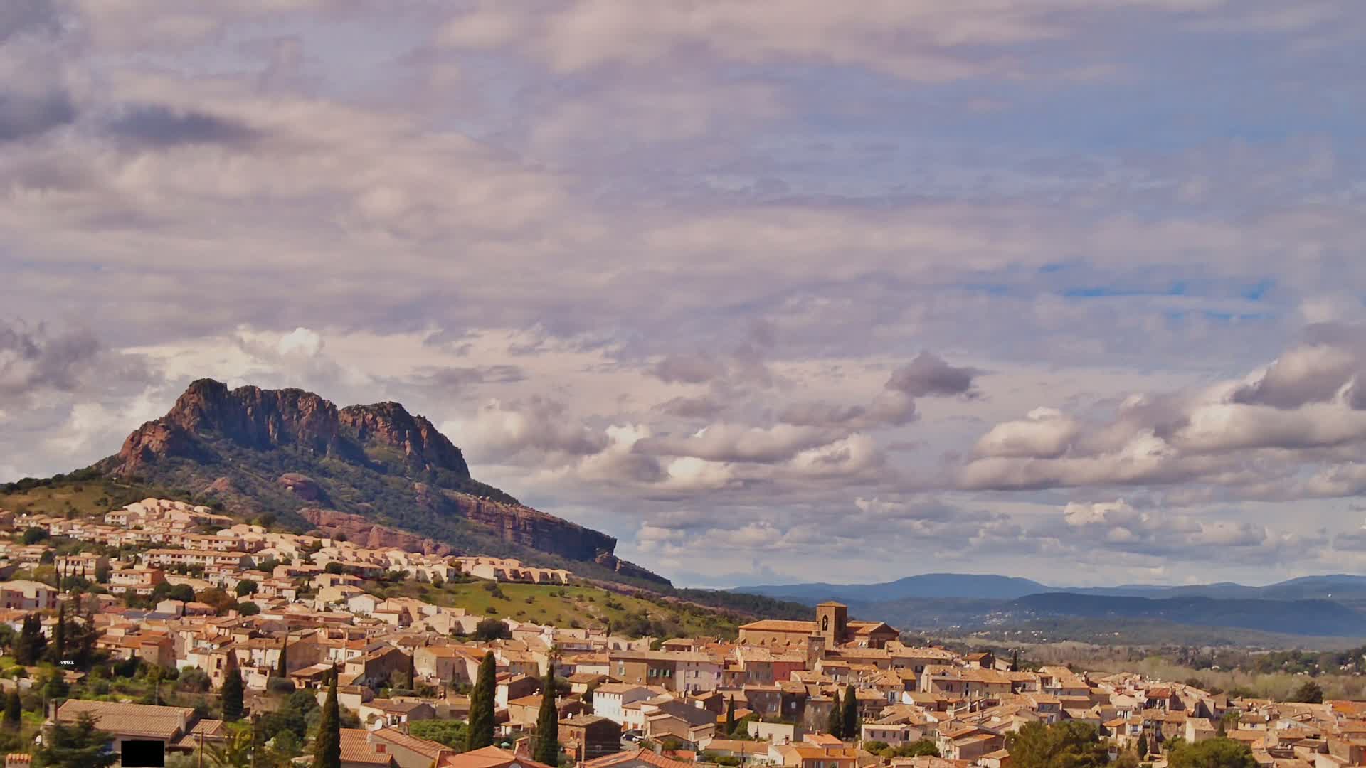 A sprawling village of tan-colored buildings nestles against a large, rocky hill under a partly cloudy sky.