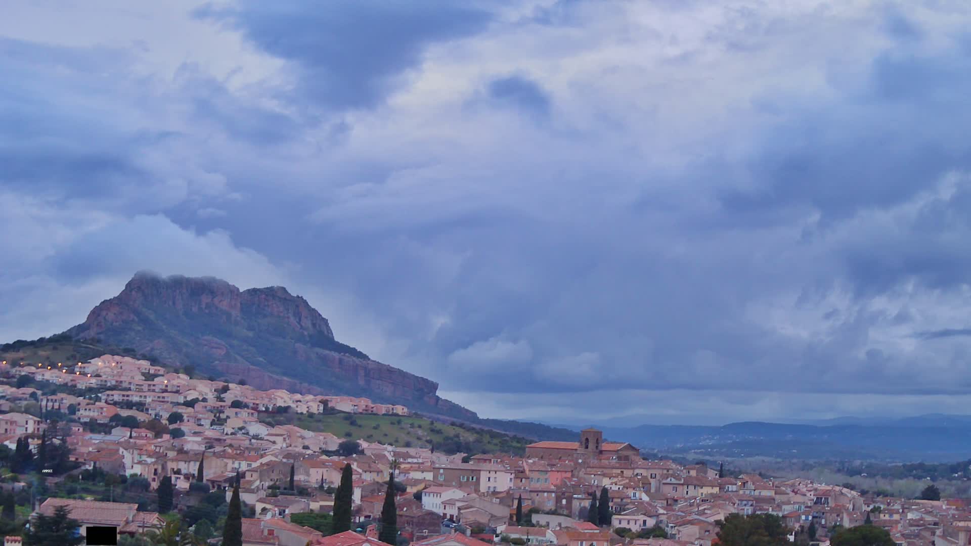 A sprawling town of light brown buildings nestles against a large, rocky hill under a mostly cloudy, overcast sky.