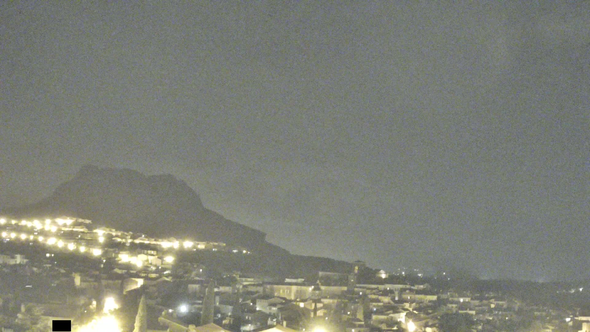 A nighttime view of a town nestled at the base of a large, dark hill under a hazy sky.