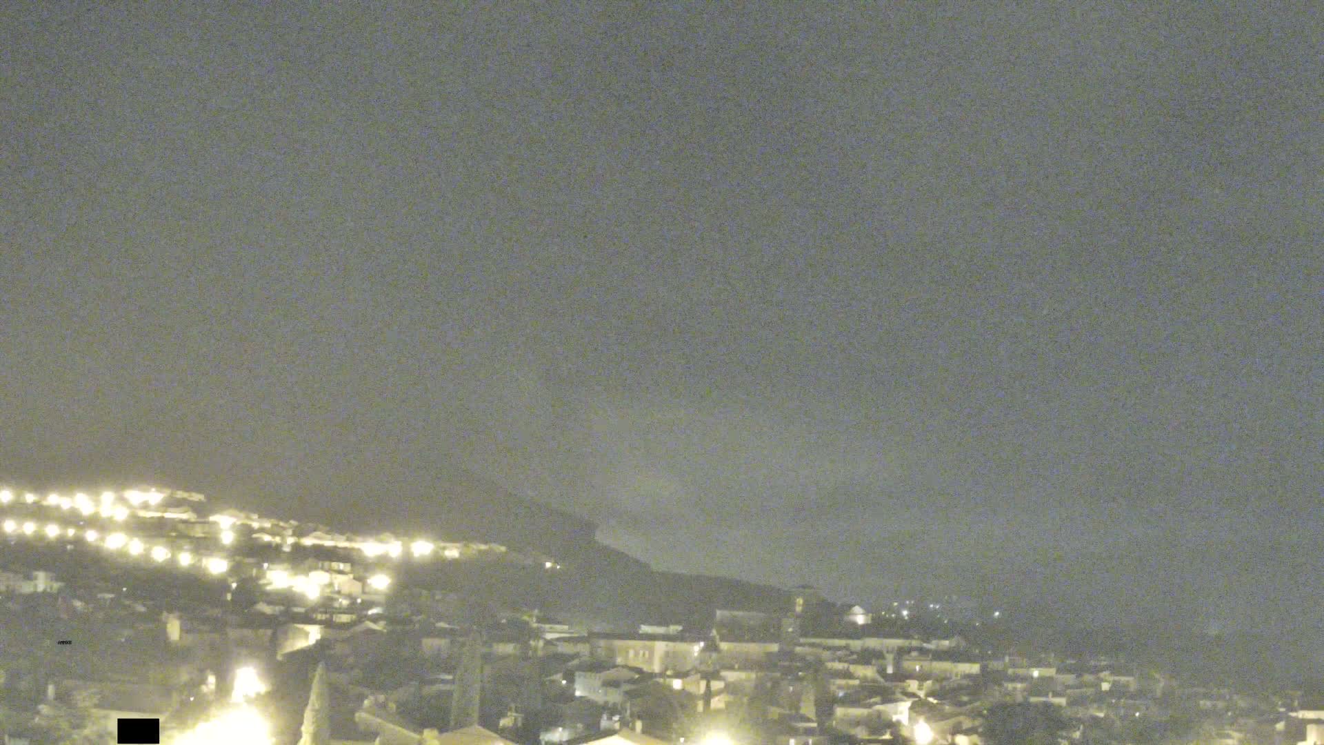 A nighttime view of a town nestled against a hill under a hazy sky.