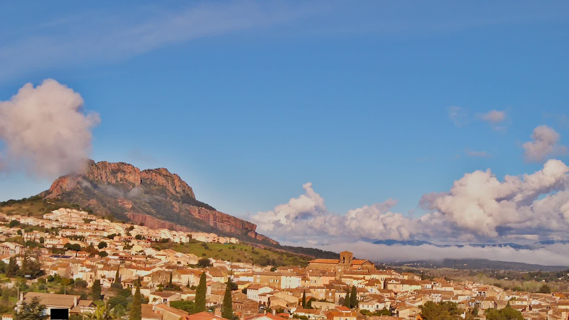 A small town of tan and brown buildings nestles at the base of a rocky, reddish-brown hill under a mostly clear blue sky with fluffy white clouds.