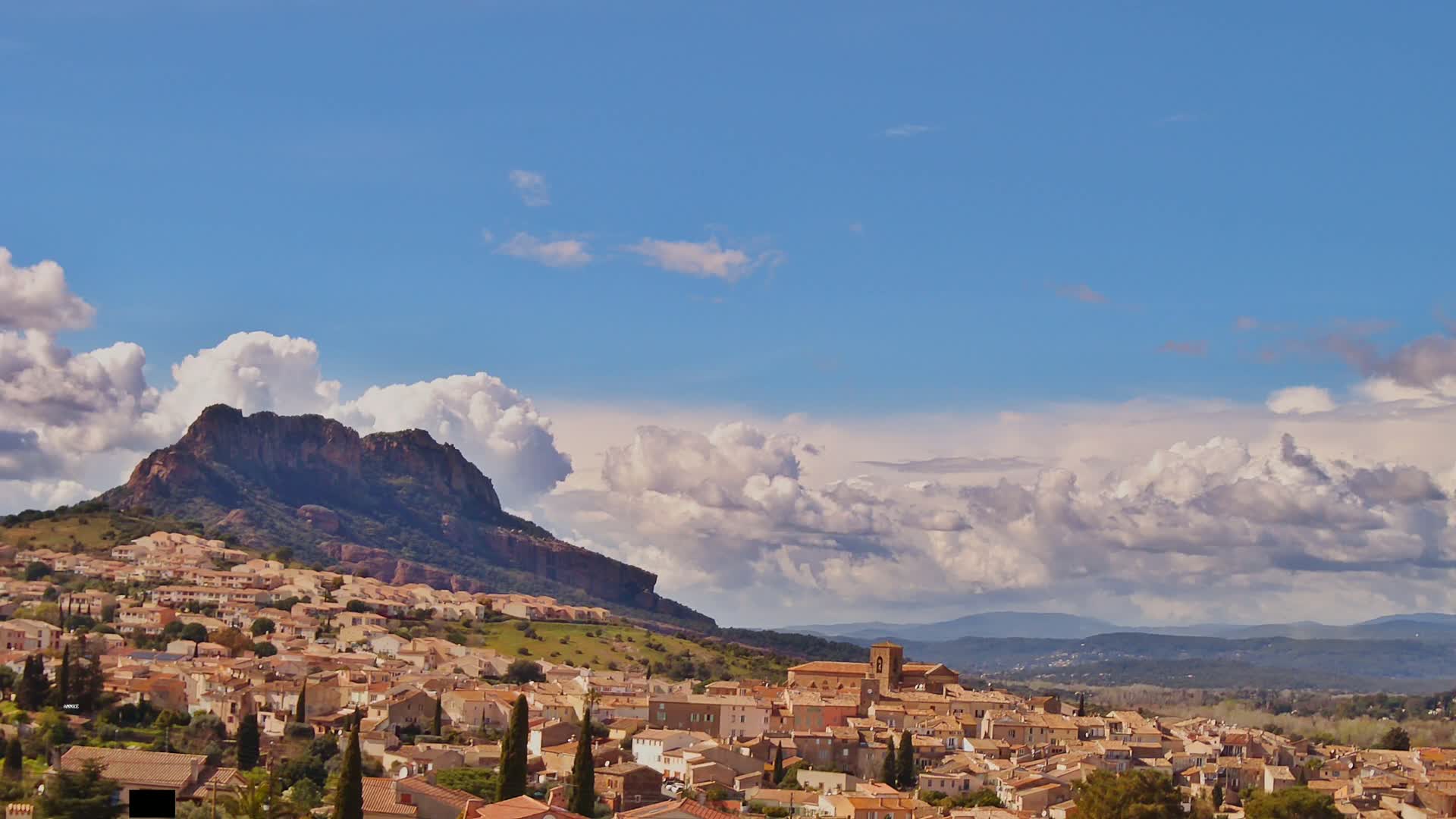 A small town of tan-colored buildings nestled against a rocky, tree-covered mountain under a partly cloudy blue sky.