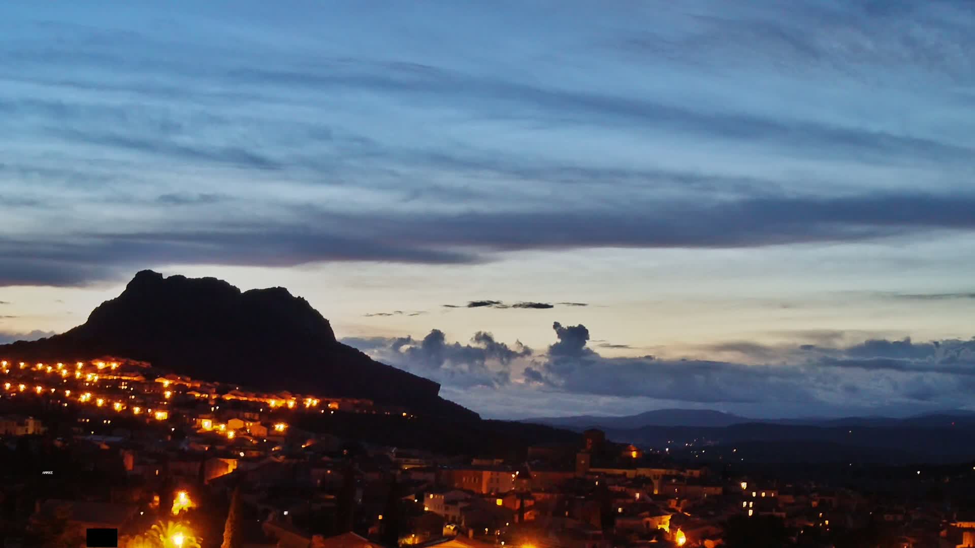 A town nestled at the base of a large, dark mountain is illuminated at night under a mostly cloudy, twilight sky.