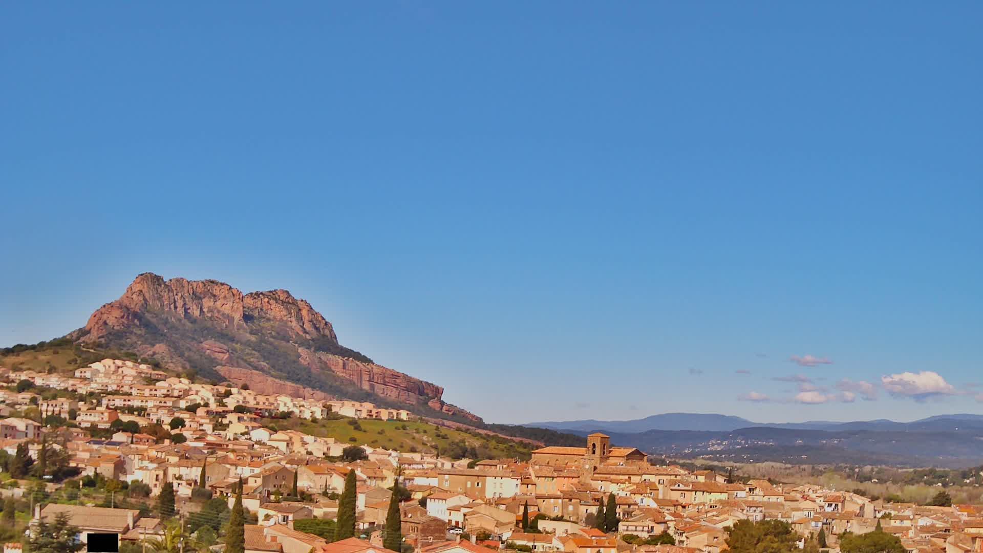 A small town of tan and brown buildings nestles on a hillside beneath a large, reddish-brown rock formation, all under a clear blue sky with a few small clouds.