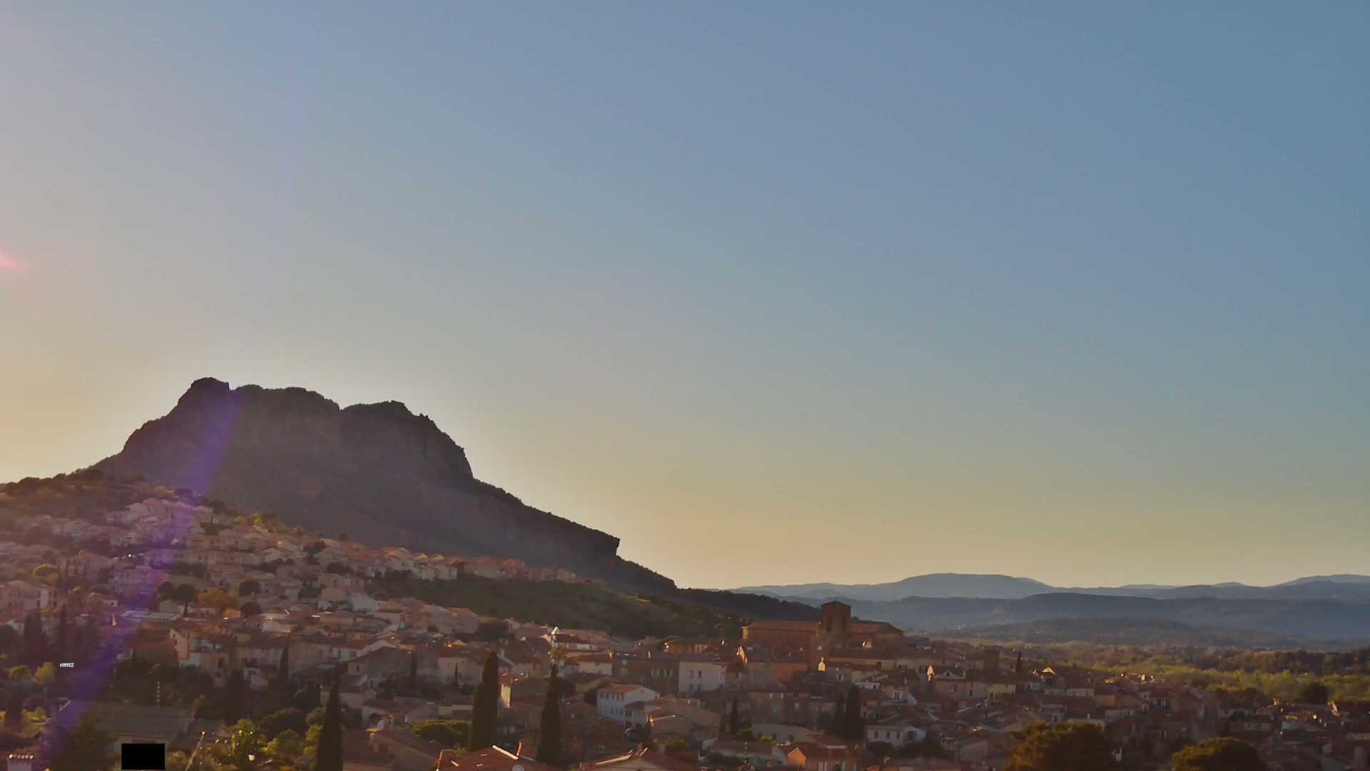 A sunlit town nestled at the base of a large, dark mountain, with distant hills visible under a clear, pale sky.
