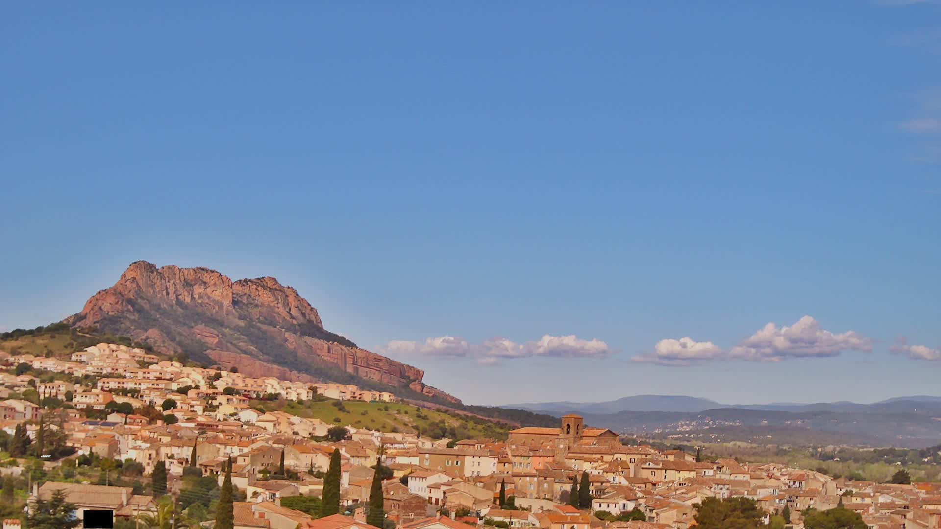 A small town of tan and brown buildings nestles against a large, reddish-brown mountain under a mostly clear blue sky with a few small, fluffy clouds.