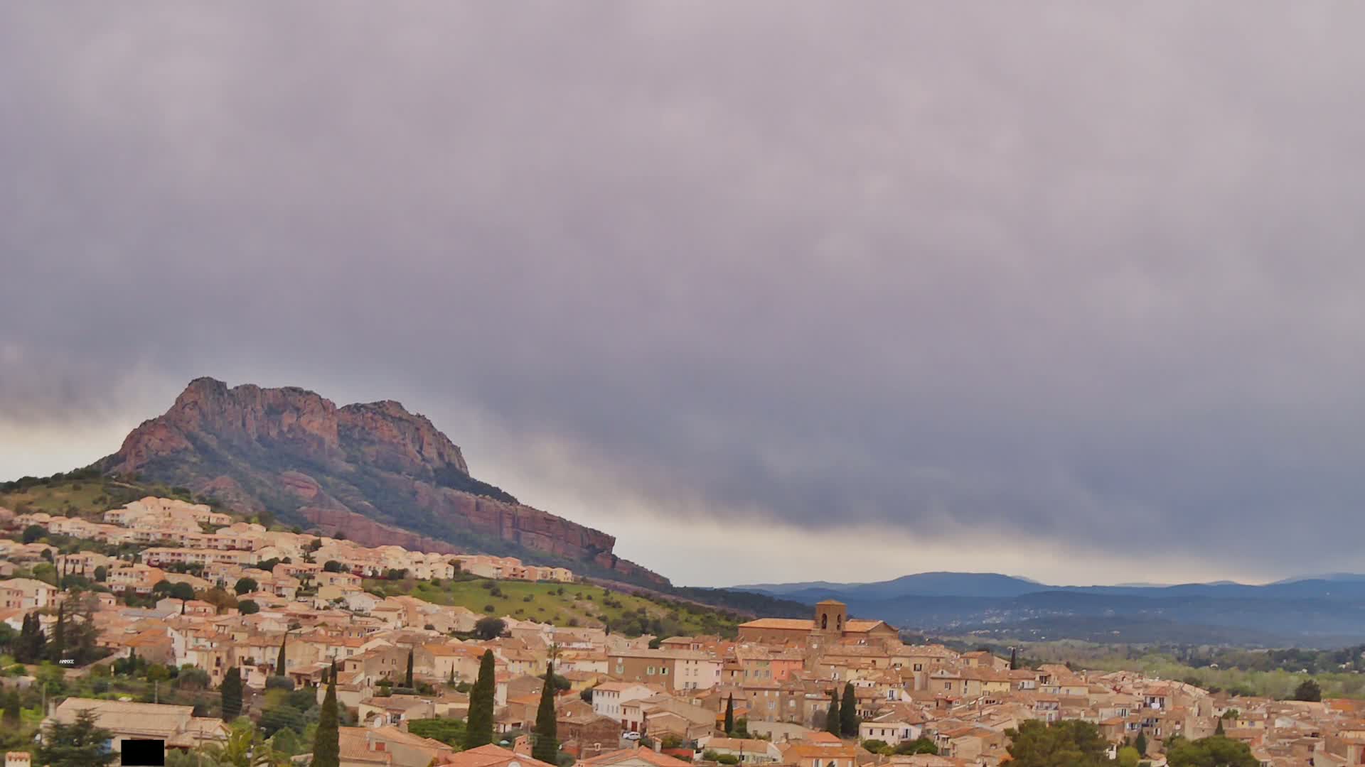 A hillside town with tan buildings nestles beneath a large, rocky mountain under a cloudy, overcast sky.