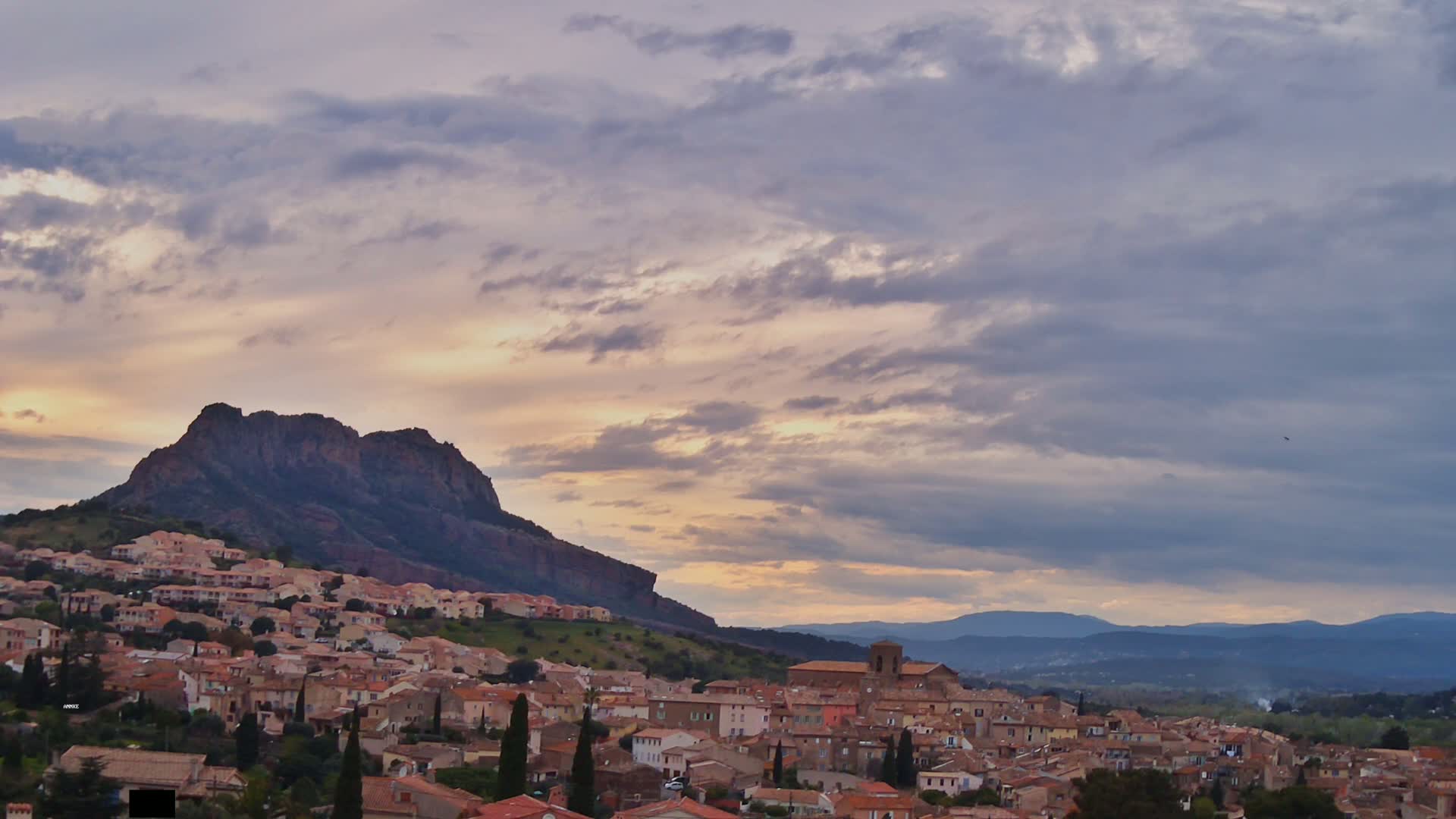 A small town nestled on a hillside below a large rock formation, under a partly cloudy sunset sky.