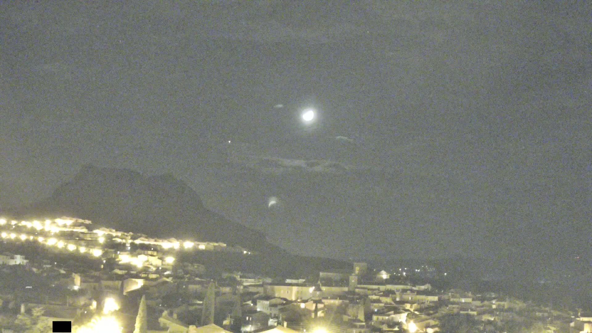 A moonlit night scene shows a hilltop town illuminated below a partly cloudy sky and a mountain in the background.