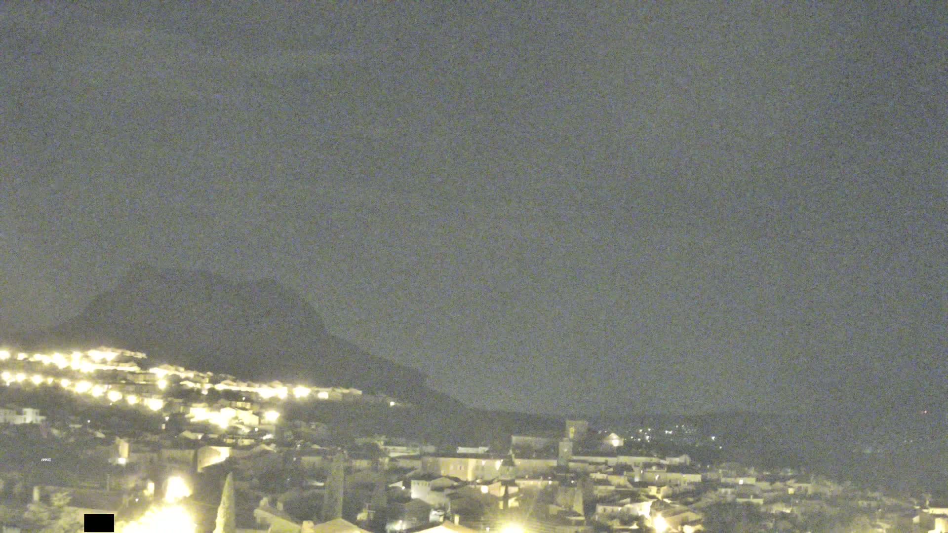 A nighttime view shows a hilltop town illuminated with scattered lights under a dark, overcast sky.