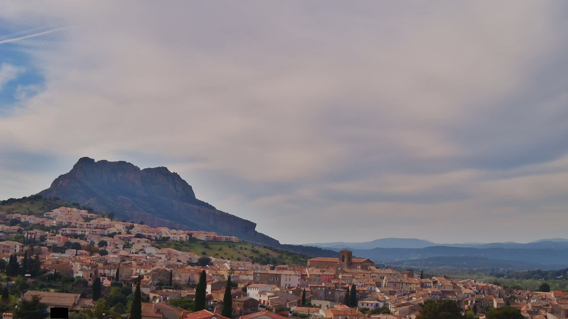 A sprawling village nestled against a large, dark mountain under a mostly cloudy sky.