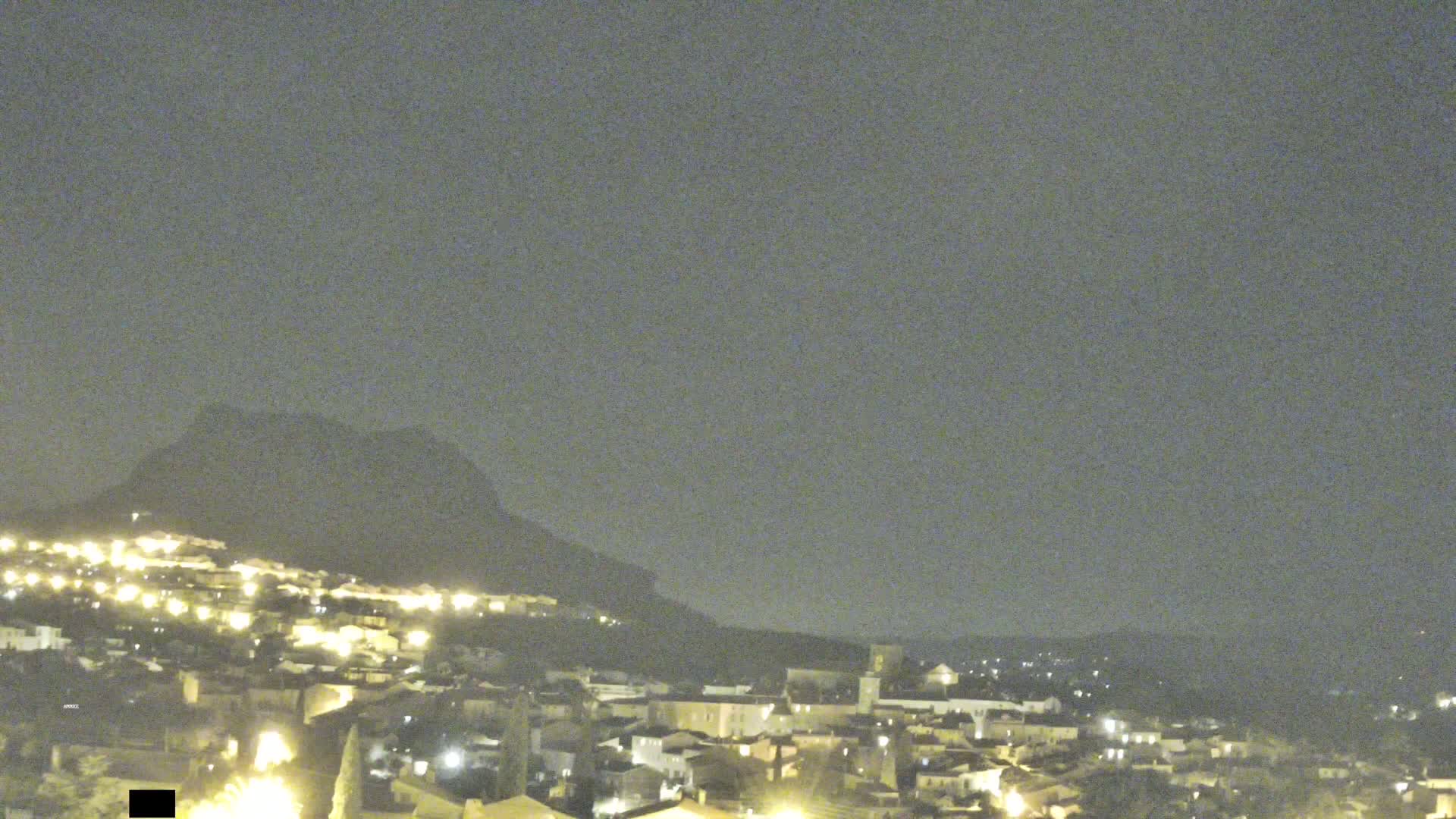 A nighttime, long shot of a town nestled against a hill, illuminated by scattered lights under a cloudy sky.