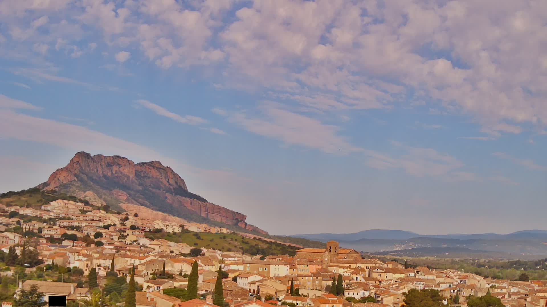 A town of tan buildings nestled on a hillside below a large reddish-brown mountain under a mostly clear sky with scattered clouds.
