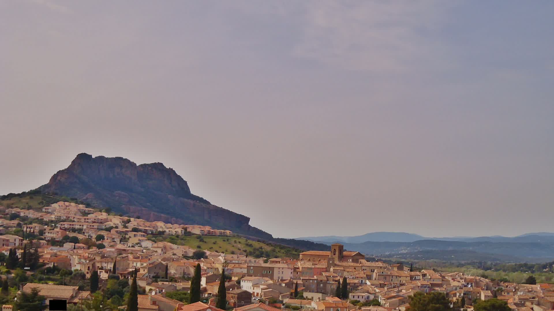 A sprawling village of tan and brown buildings nestles against a large, dark mountain under a hazy, light-purple sky.