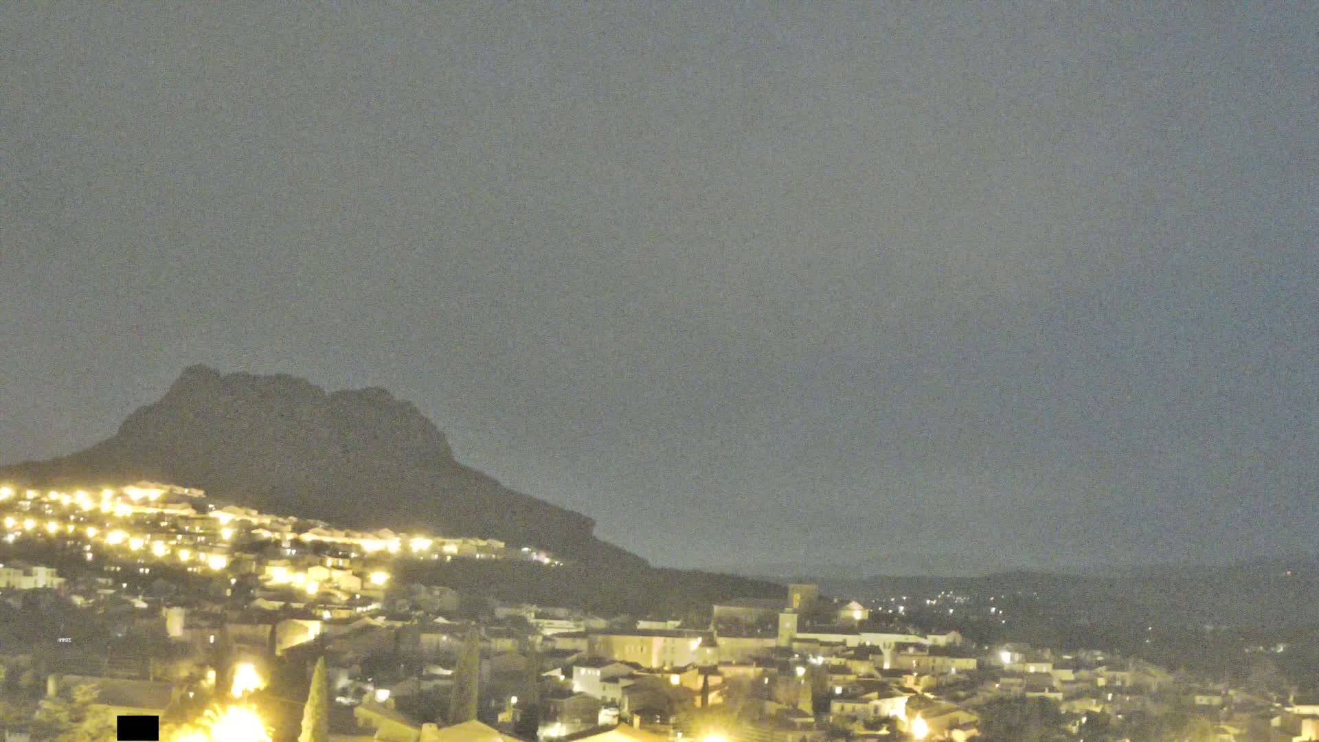 A nighttime view of a town nestled at the base of a dark mountain under a hazy sky.