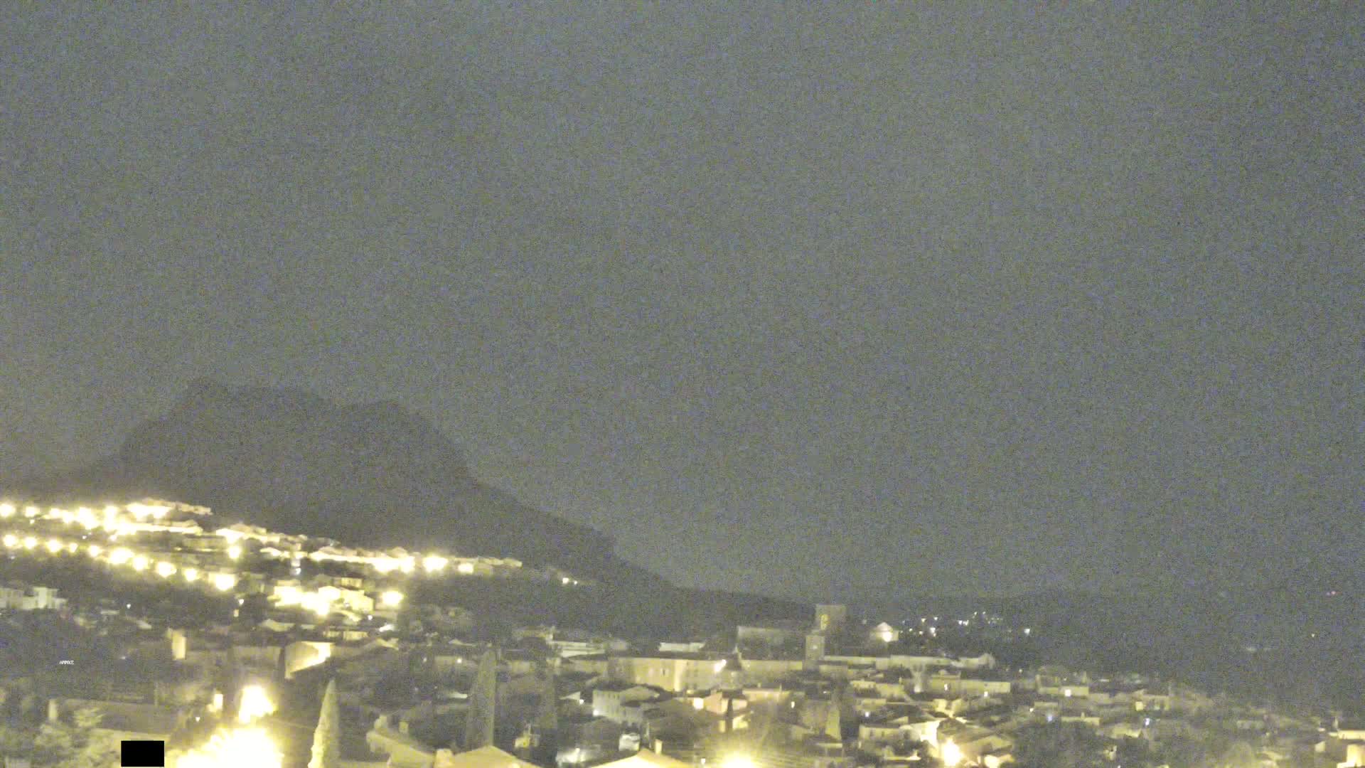 A nighttime view of a town nestled against a hill, illuminated by scattered lights under a dark, overcast sky.