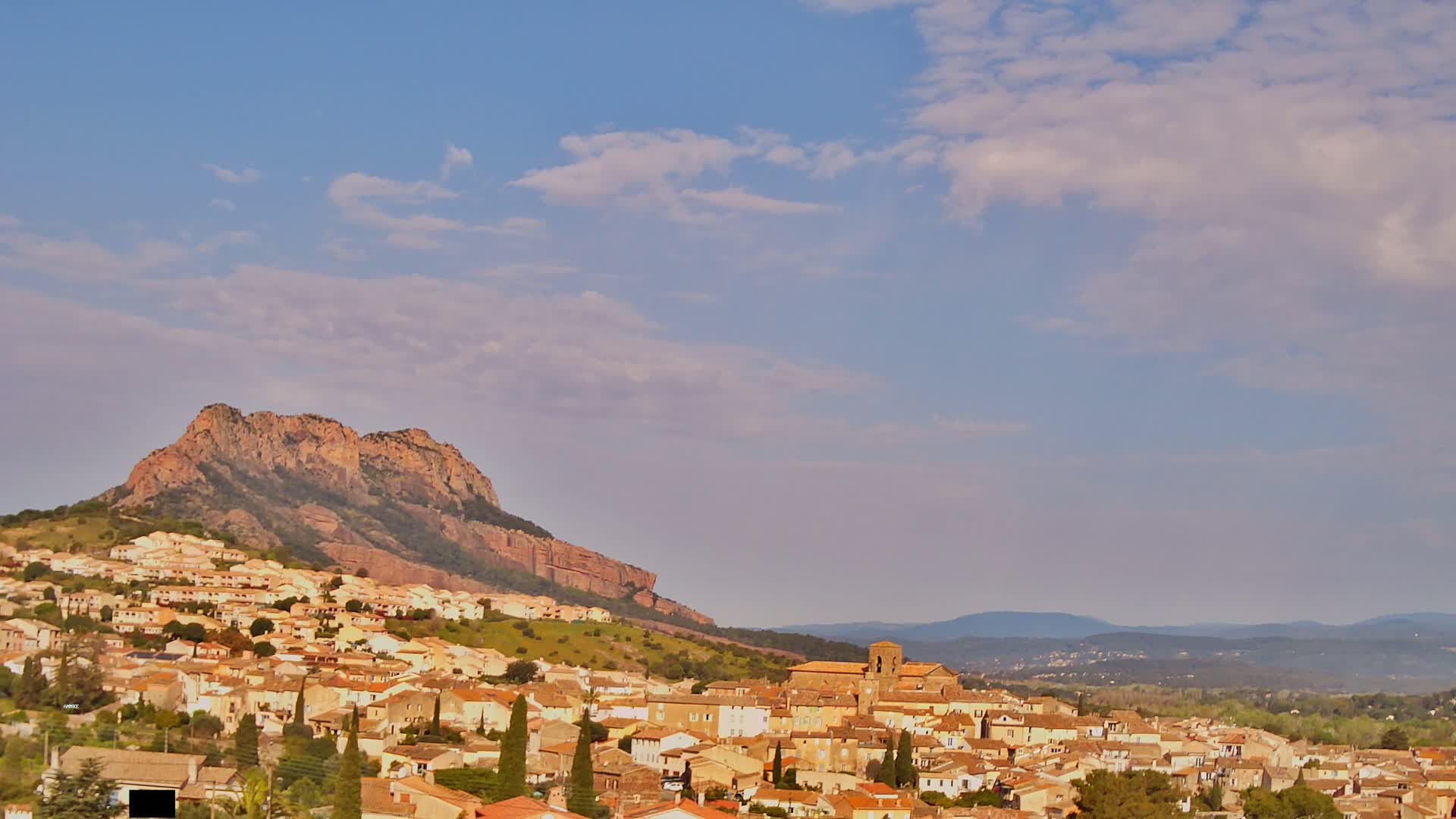 A town of tan and brown buildings nestled on a hillside below a large, reddish-brown mountain under a mostly clear, light blue sky with scattered clouds.