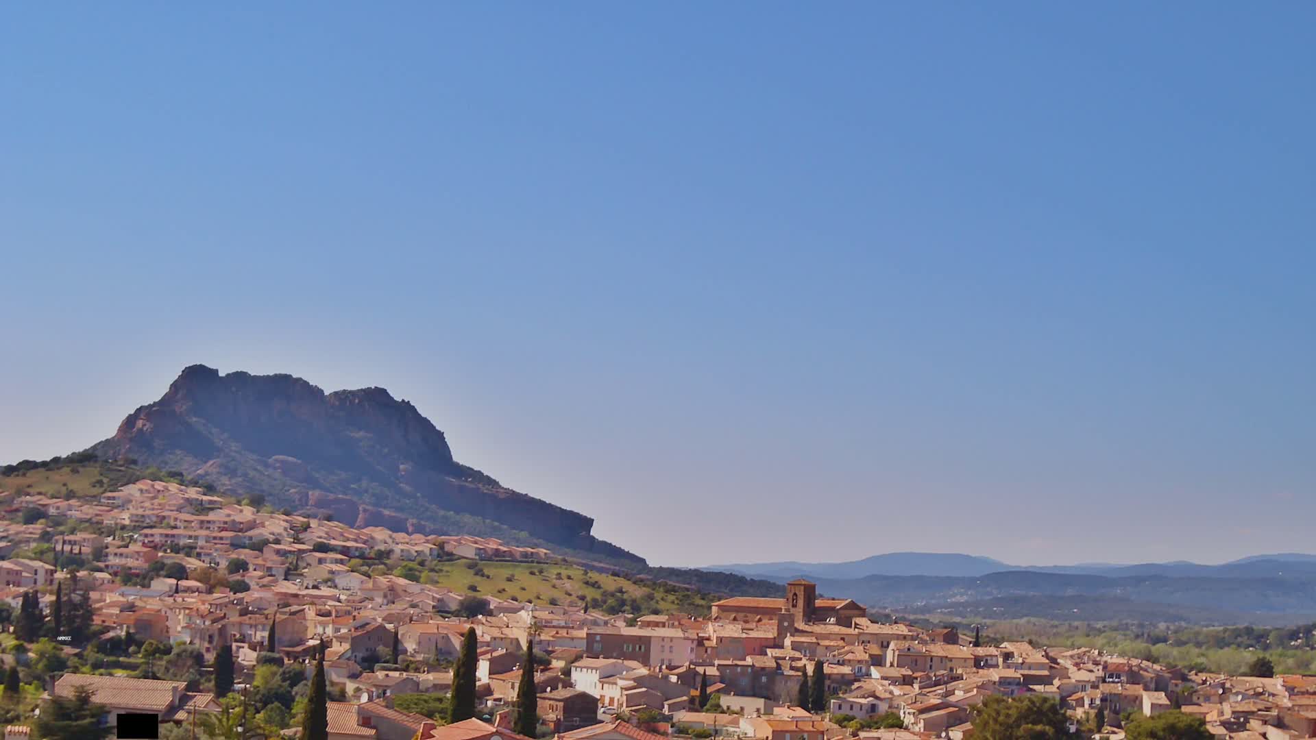 A sprawling town of tan buildings nestles at the base of a large, dark mountain under a clear blue sky.