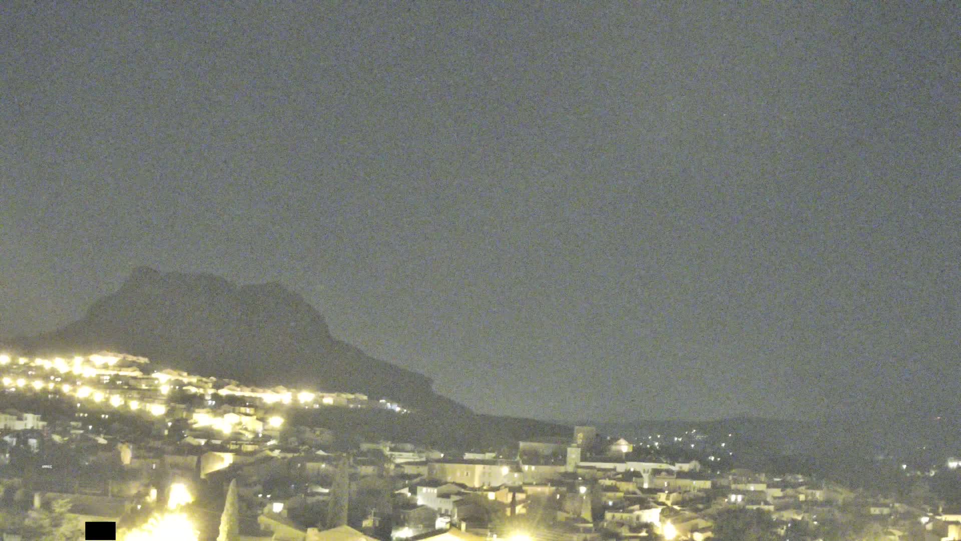 A nighttime view of a town nestled at the base of a dark mountain under a cloudy sky.