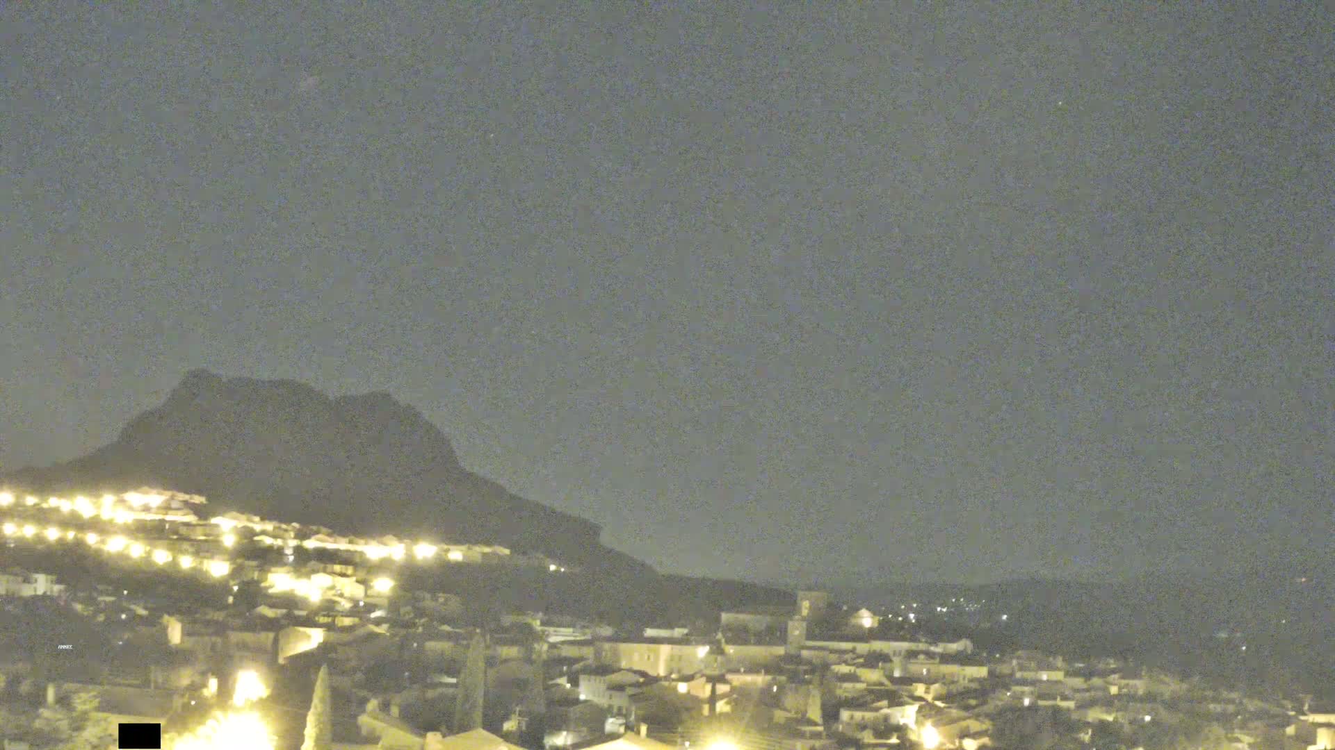A nighttime view shows a town illuminated by lights nestled against a dark mountain under a clear, dark sky.