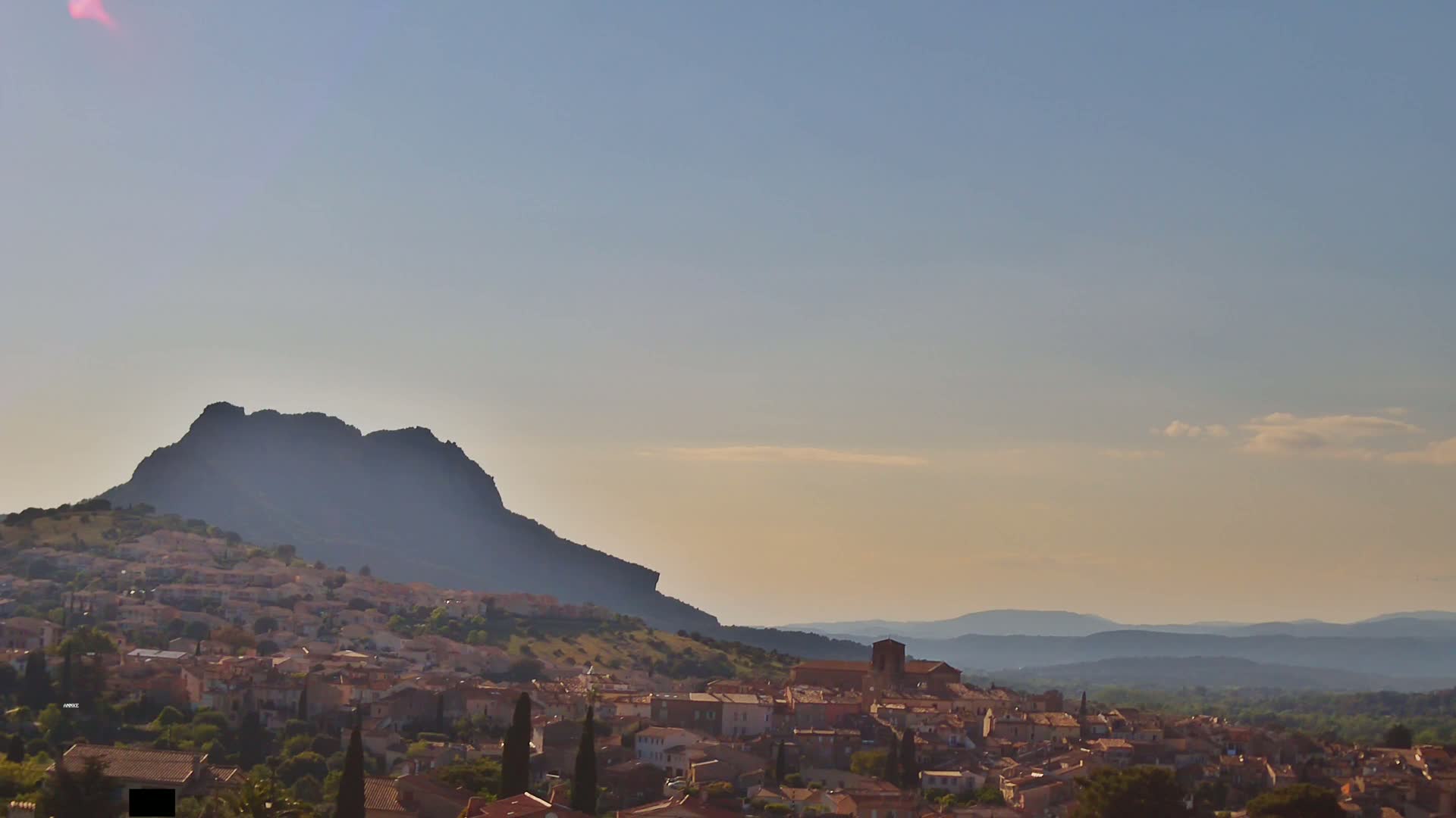 A large, dark hill rises behind a sprawling town nestled in a valley, under a clear, pale sky.