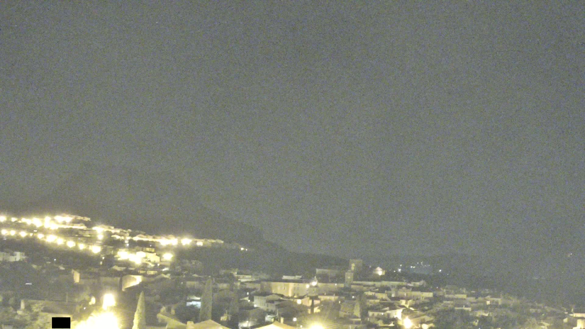 A nighttime, long shot shows a hillside town illuminated by many small, scattered lights under a dark, overcast sky.