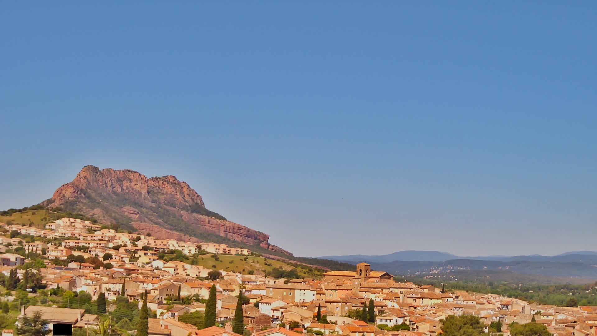 A tan-colored town sits nestled against a large, reddish-brown mountain under a clear blue sky.