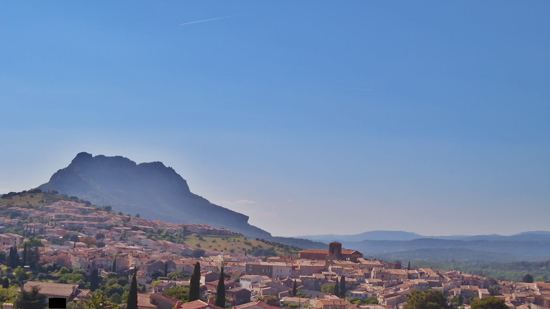 A small town with tan-colored buildings nestled on a hillside beneath a large, dark mountain under a clear blue sky.