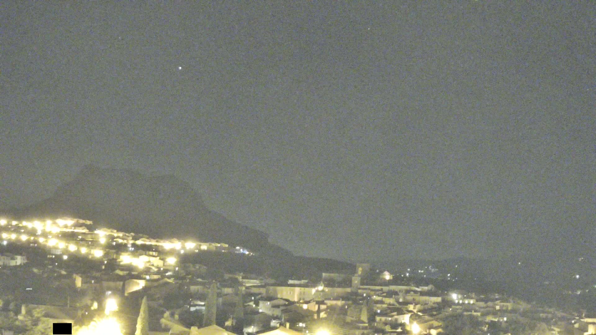A nighttime view of a town nestled at the base of a dark mountain under a clear, starry sky.