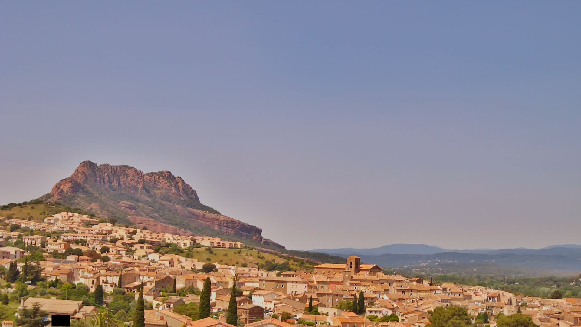 A tan-colored village is nestled at the base of a large, reddish-brown rock formation under a clear, light-blue sky.