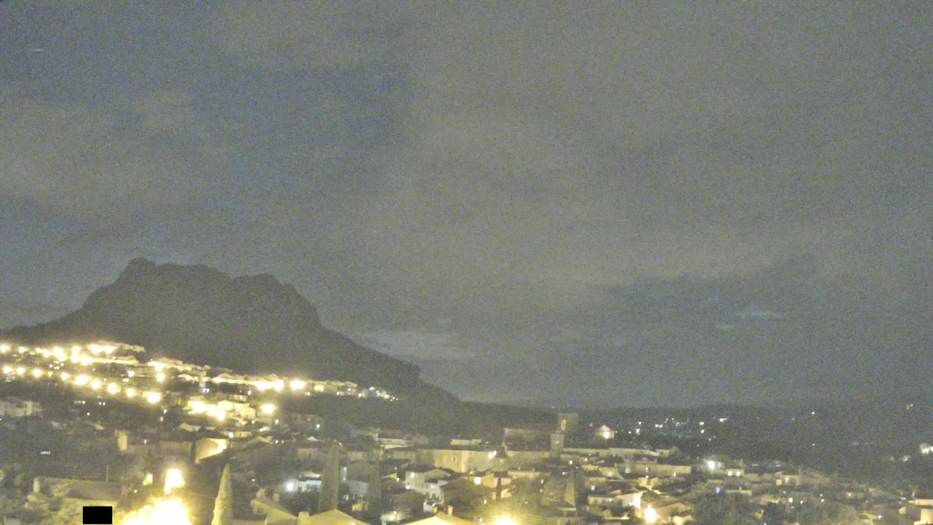 A nighttime view of a town nestled at the base of a dark mountain under a cloudy sky.