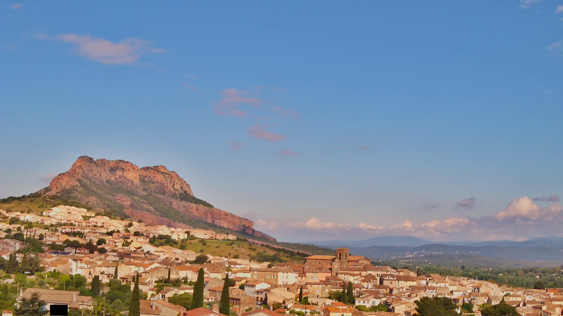 A small town with tan buildings nestled at the base of a large, reddish-brown mountain under a mostly clear, light-blue sky.