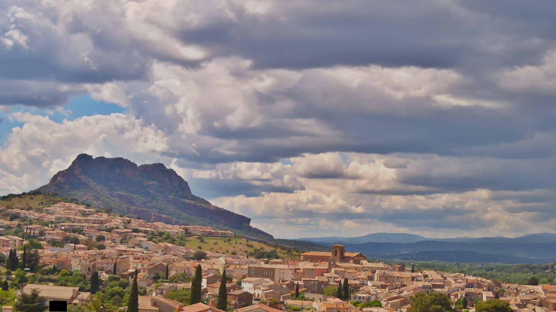A partly cloudy sky hangs over a small town nestled at the base of a large, rocky hill, with distant mountains visible on the horizon.