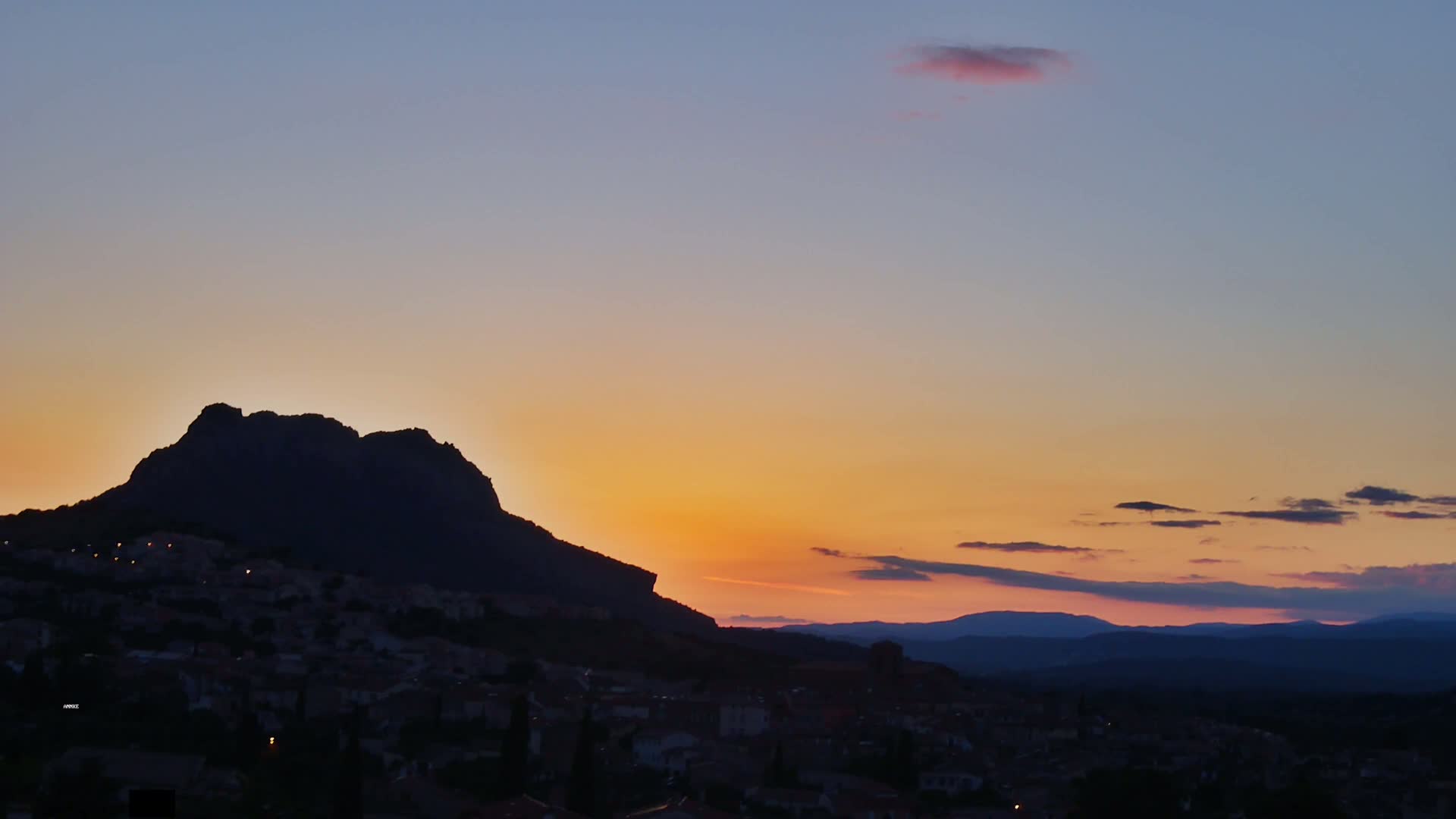 A large, dark mountain silhouetted against a sunset sky, with a town nestled at its base.