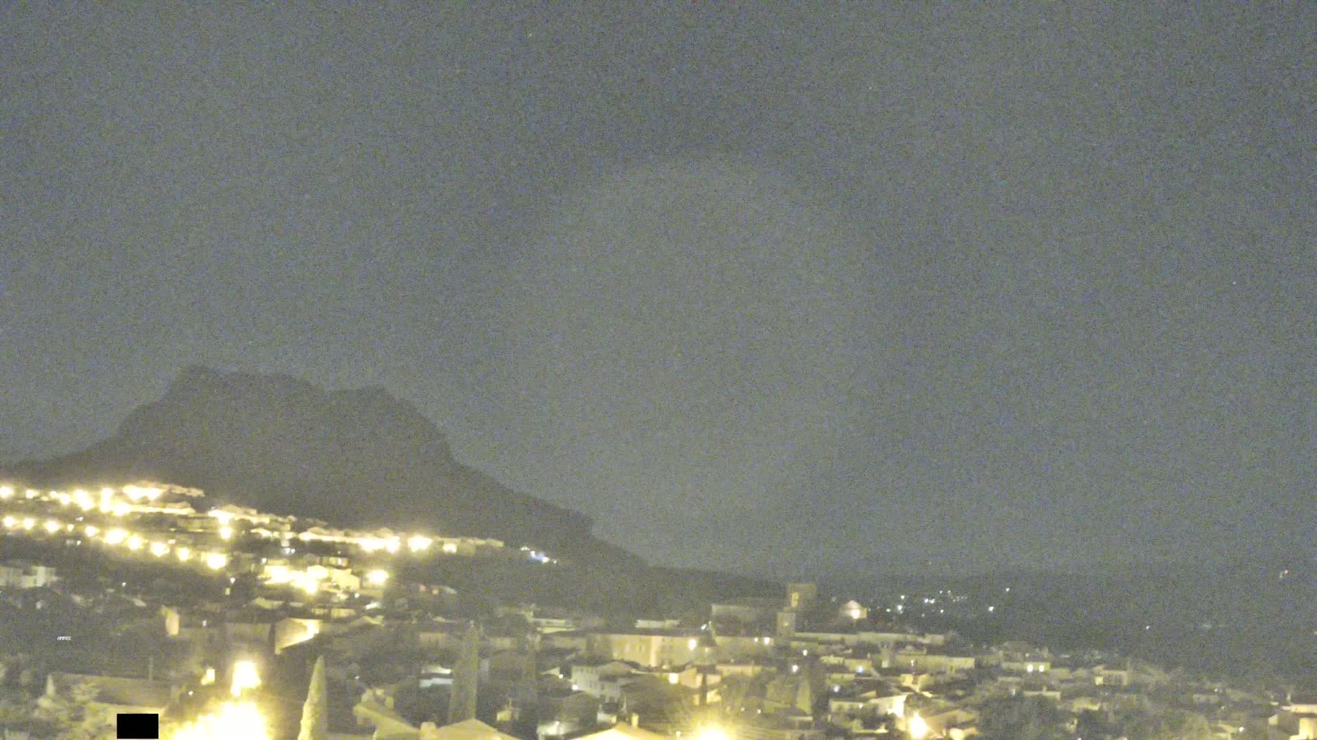 A nighttime view of a town nestled at the base of a dark mountain under a cloudy sky.