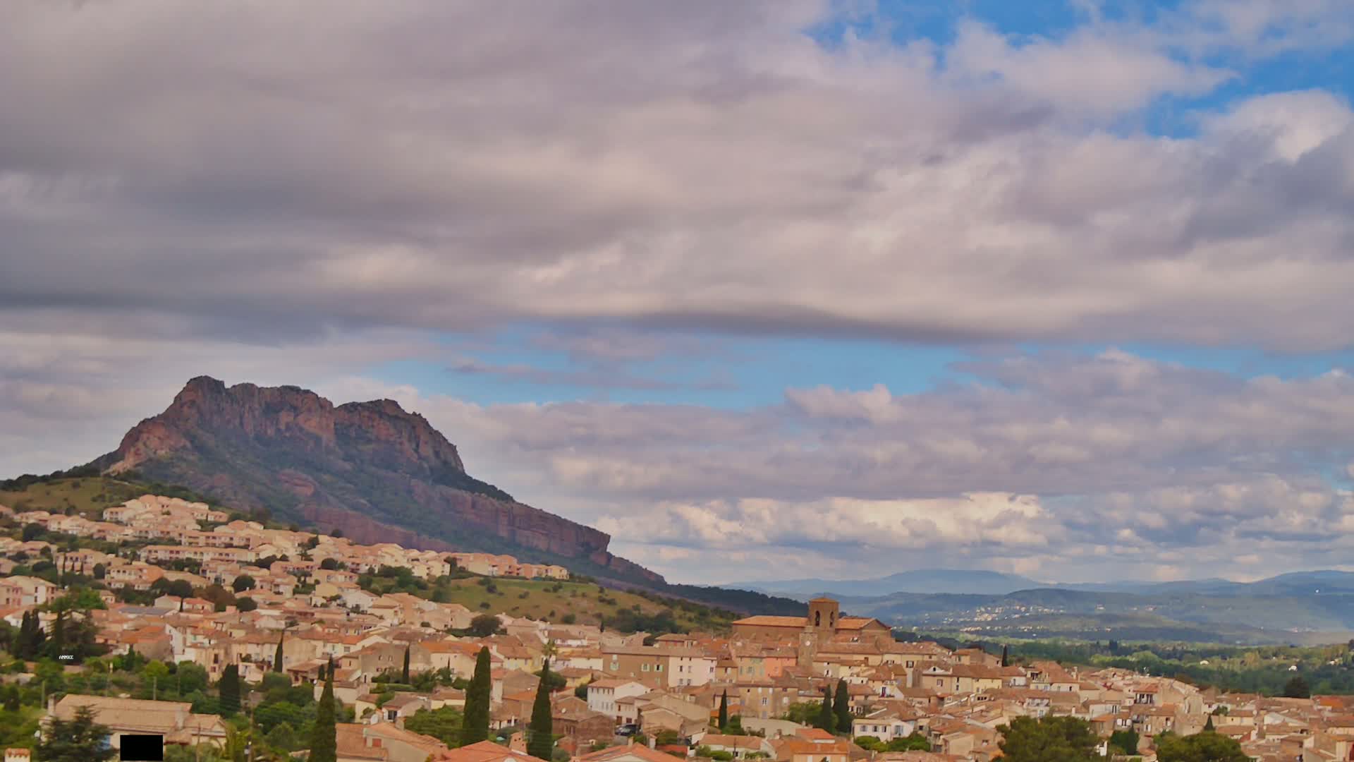 A sprawling town of tan-colored buildings nestles at the base of a large, dark-red mountain under a partly cloudy sky.