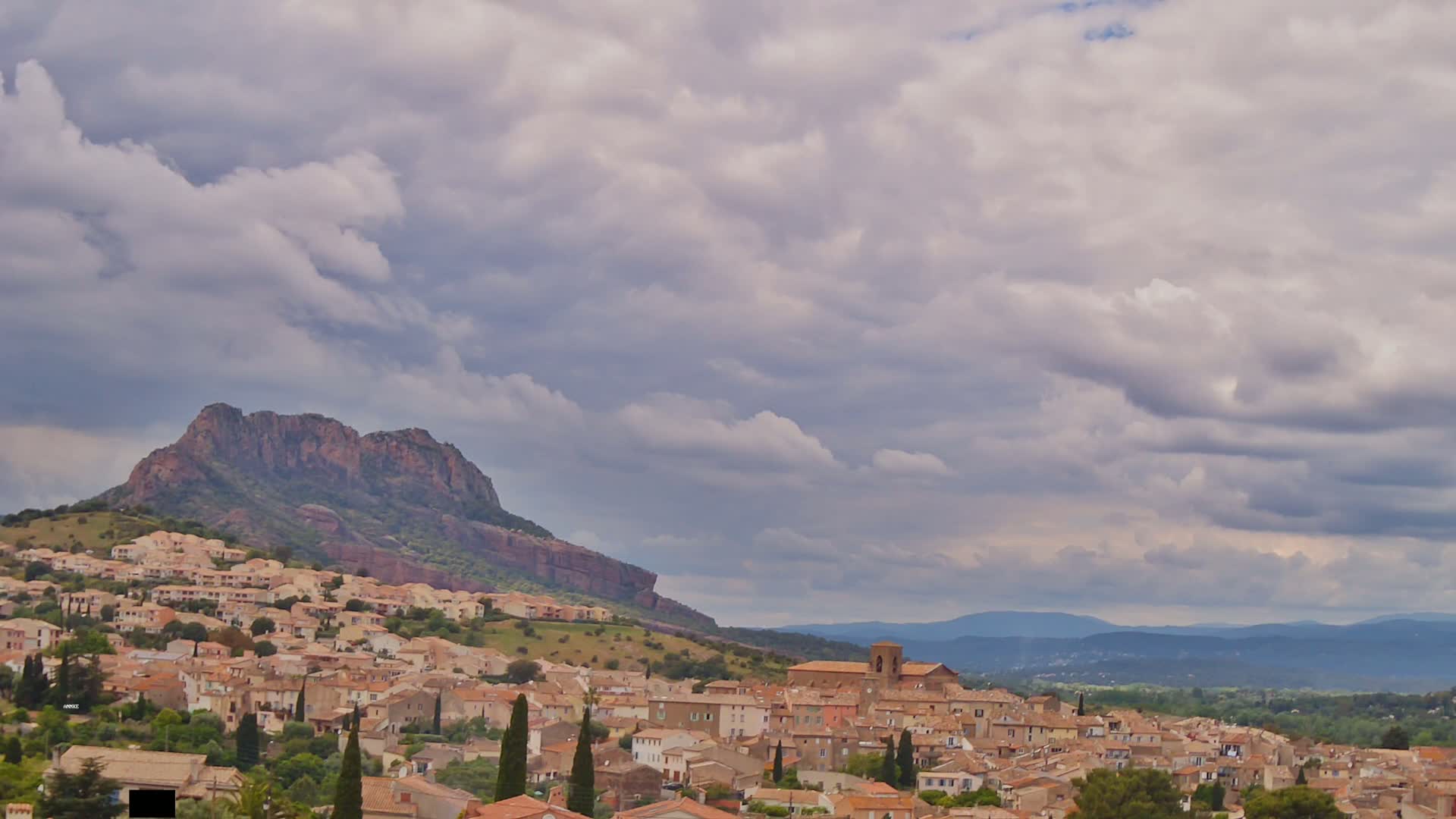 A sprawling village of tan, low buildings nestled against a large, rocky hill under a cloudy sky.