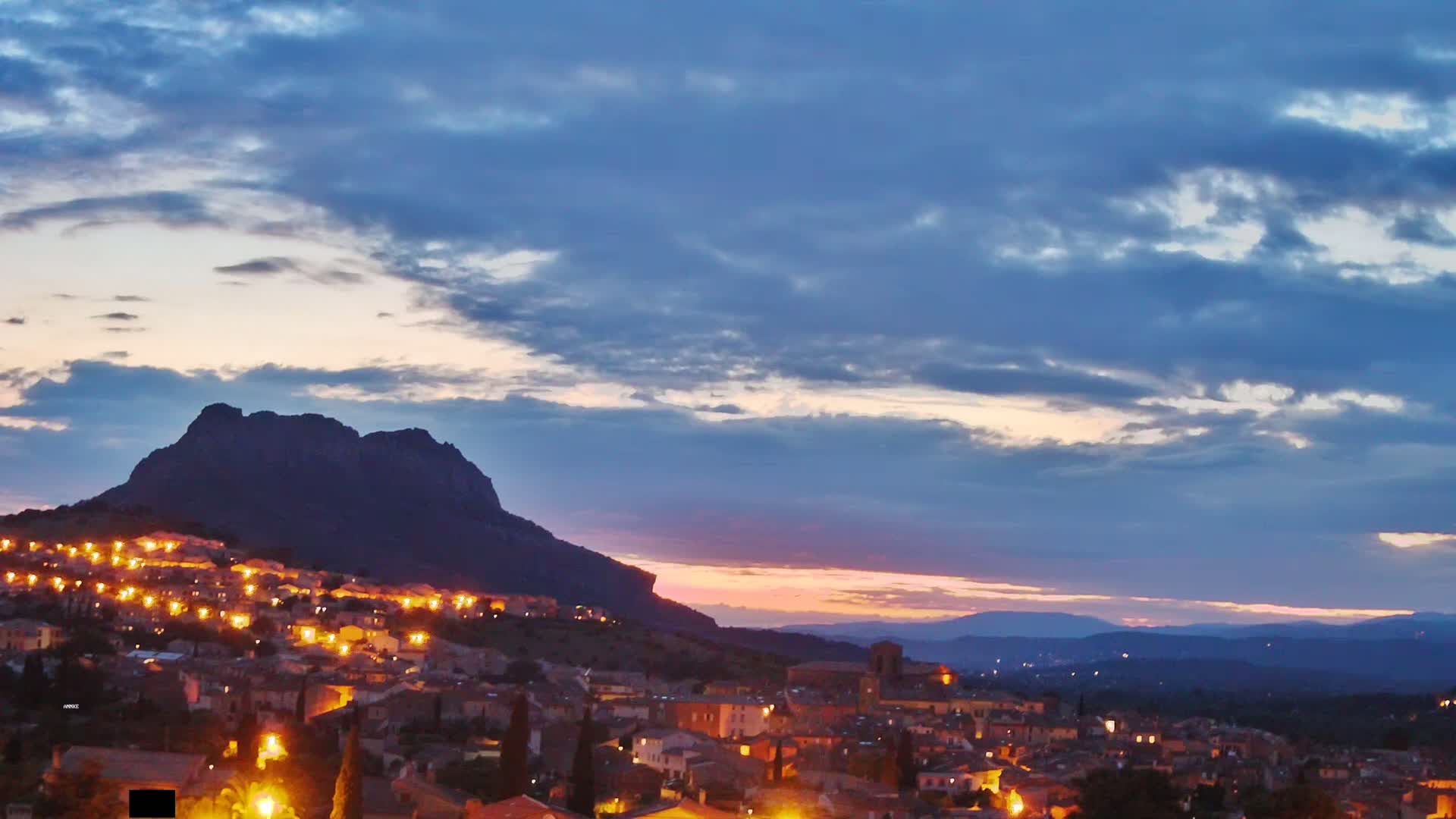 A town illuminated at twilight is nestled at the foot of a large, dark mountain under a partly cloudy sky with a hint of pink on the horizon.