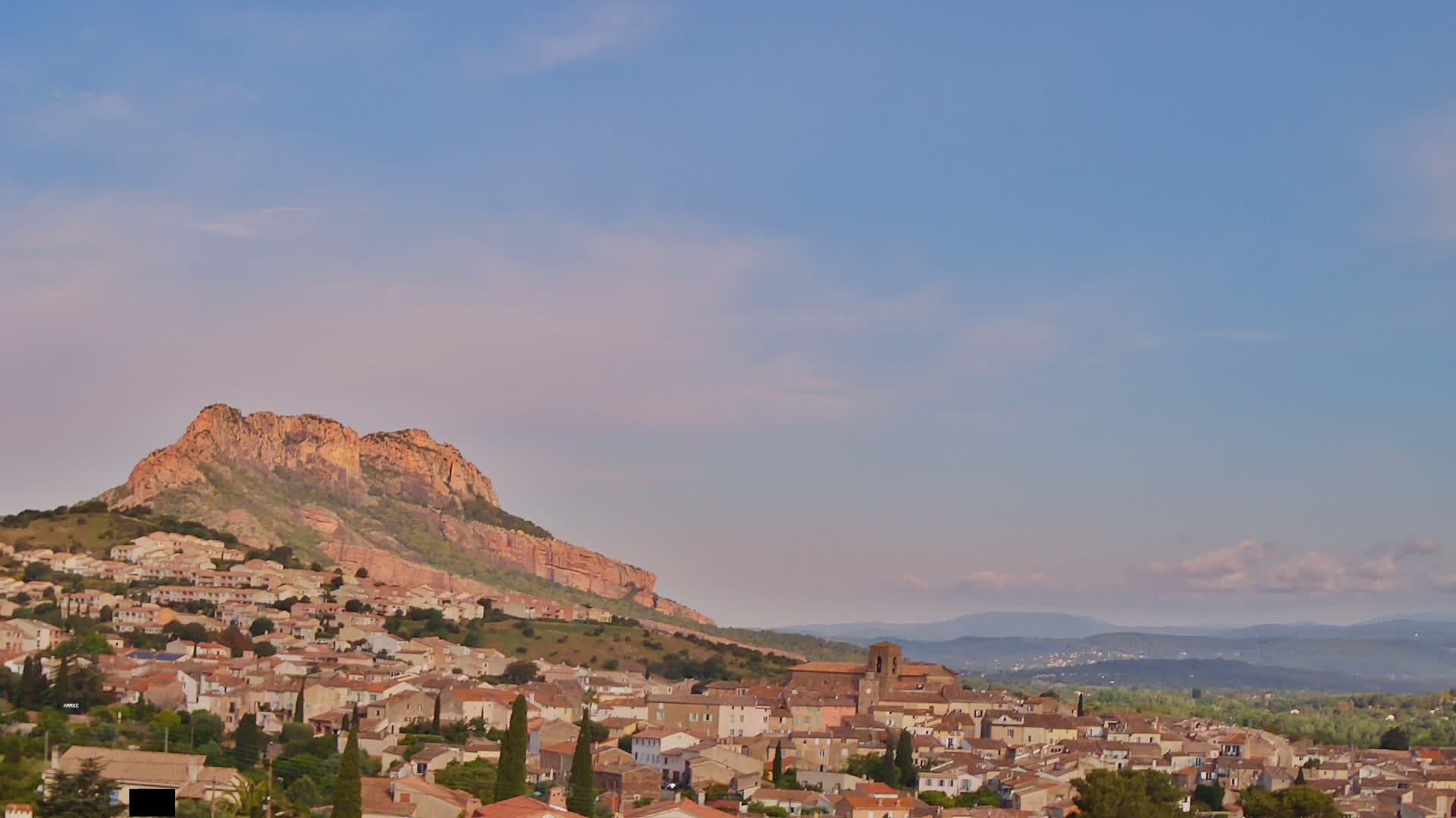 A sprawling town of low, tan buildings nestles against a large, reddish-brown hill under a pale blue, mostly clear sky.