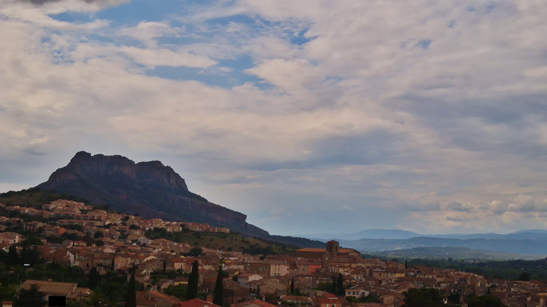 A town of tan and brown buildings sits on a hillside below a dark, rocky mountain under a partly cloudy sky.