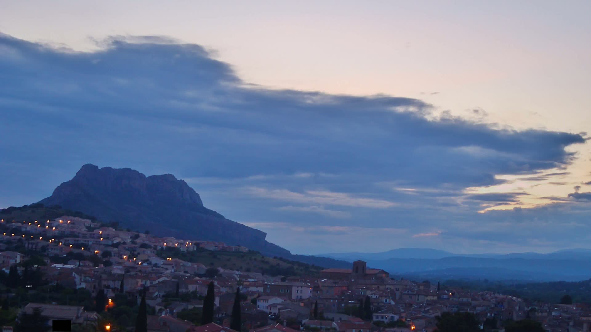 A hilltop town with illuminated buildings sits beneath a partly cloudy twilight sky, backed by a large, dark mountain and a range of distant mountains.
