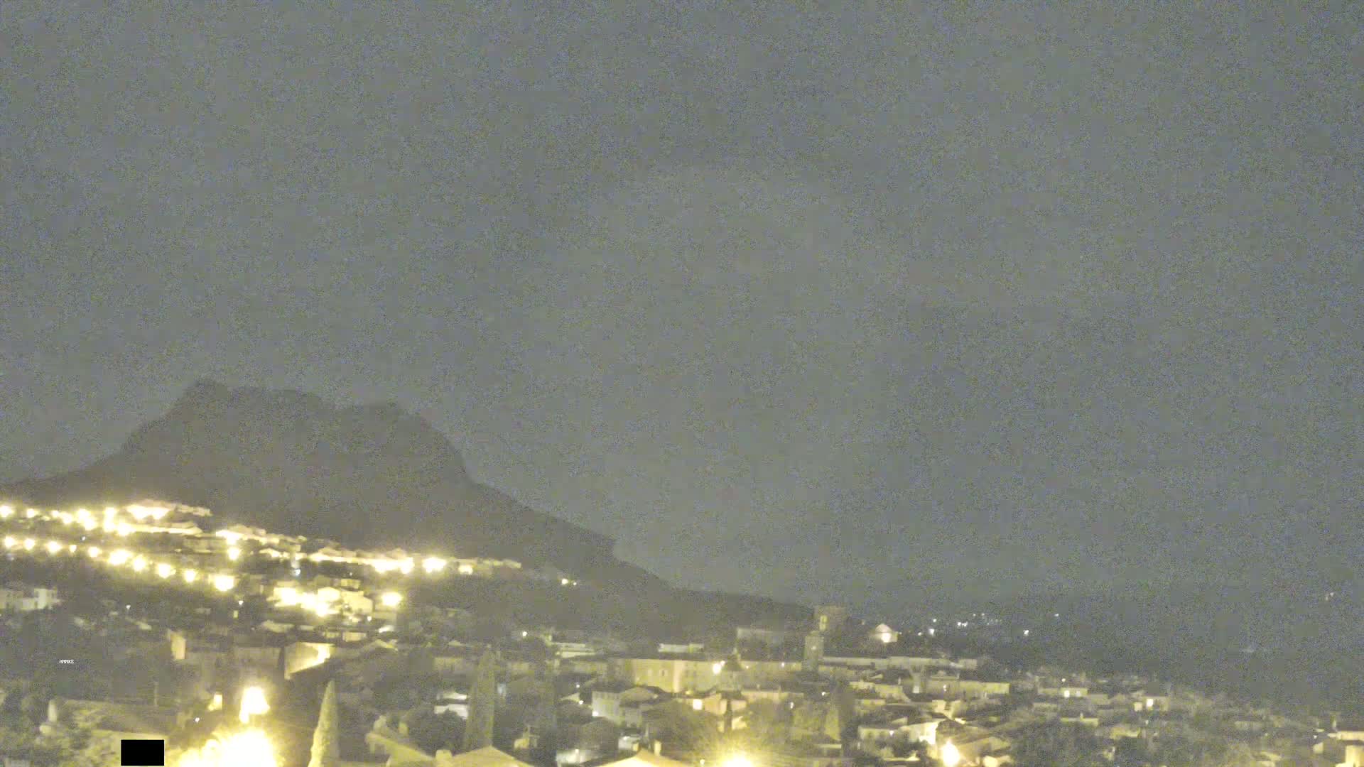 A nighttime view shows a village illuminated by lights nestled at the base of a dark mountain under a cloudy sky.