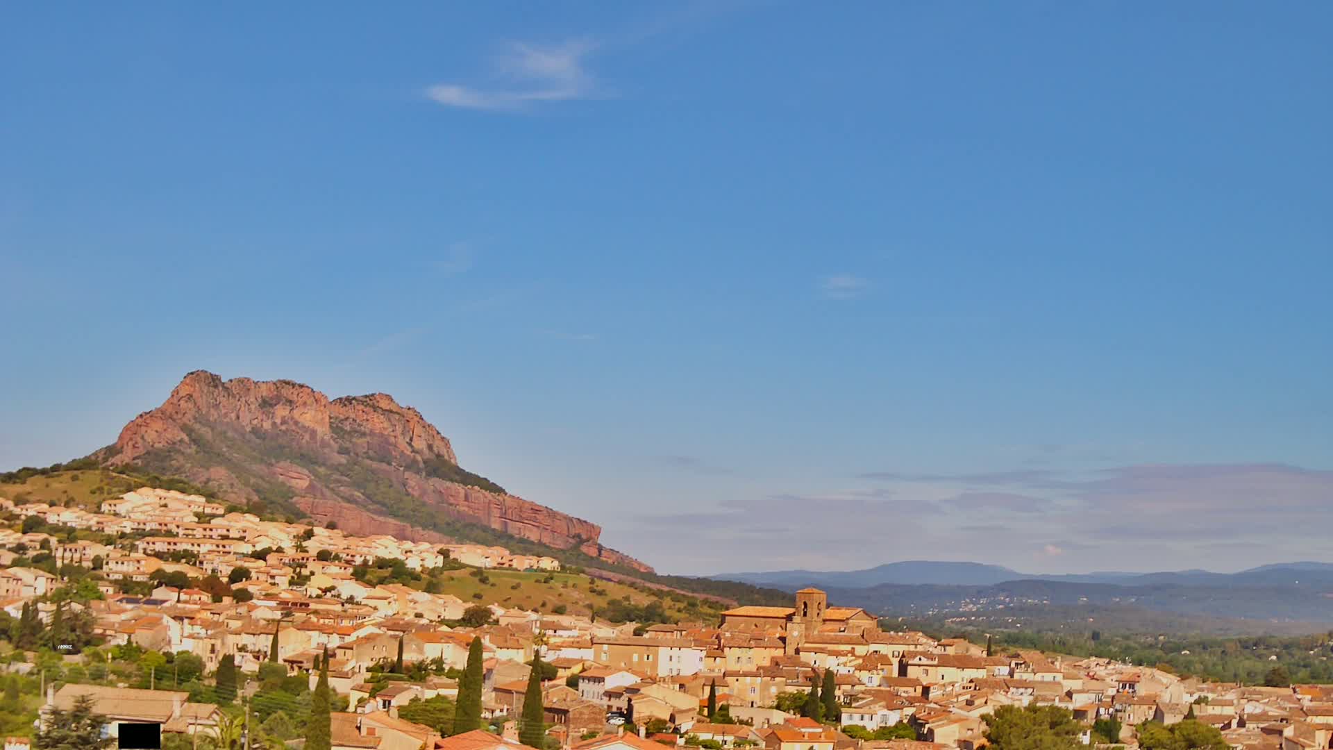 A small town of tan and orange buildings sprawls across a hillside beneath a large reddish-brown mountain under a clear blue sky.