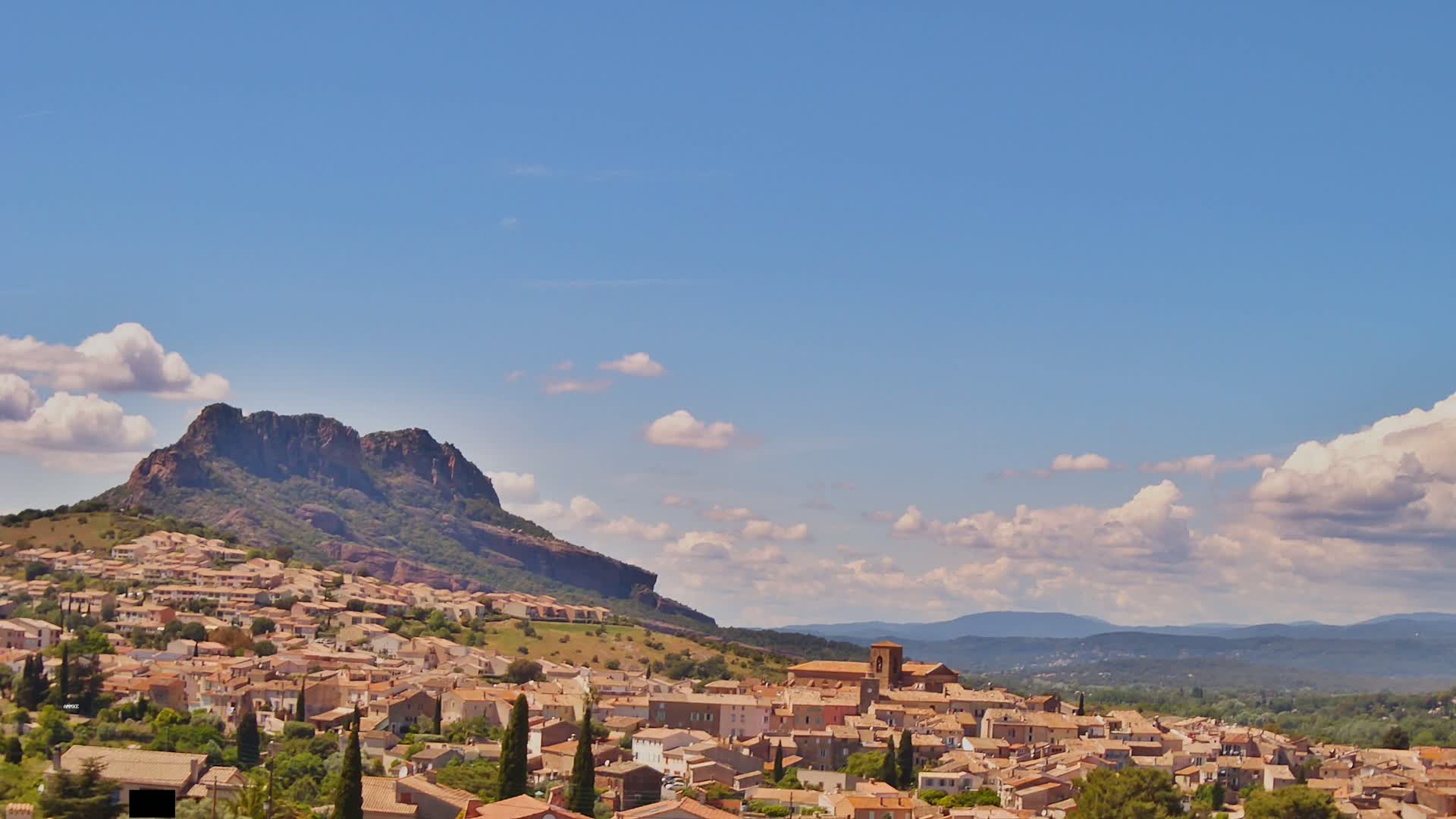 A sprawling town of light brown buildings nestles at the base of a large, dark-green, rocky hill under a mostly sunny sky with scattered cumulus clouds.