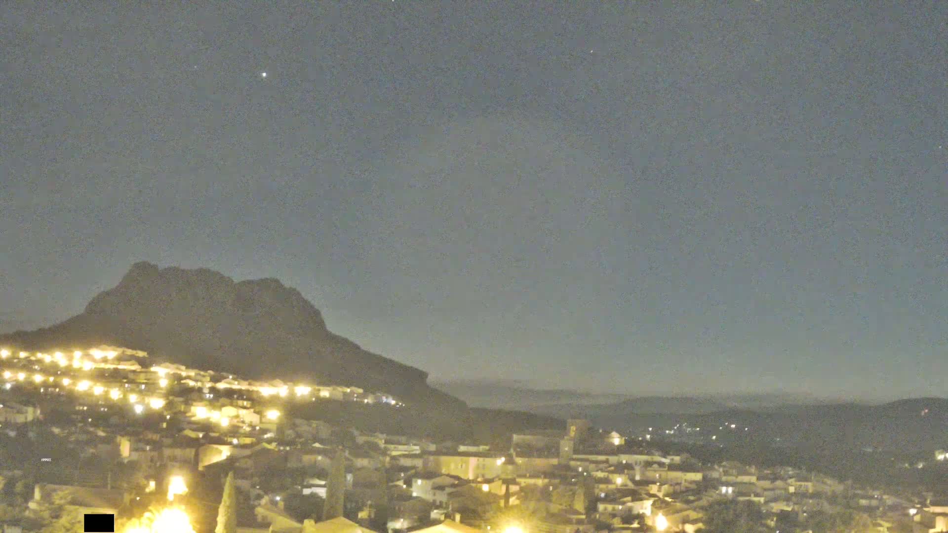 A nighttime view shows a town illuminated with lights nestled at the base of a dark mountain under a clear, slightly hazy sky.