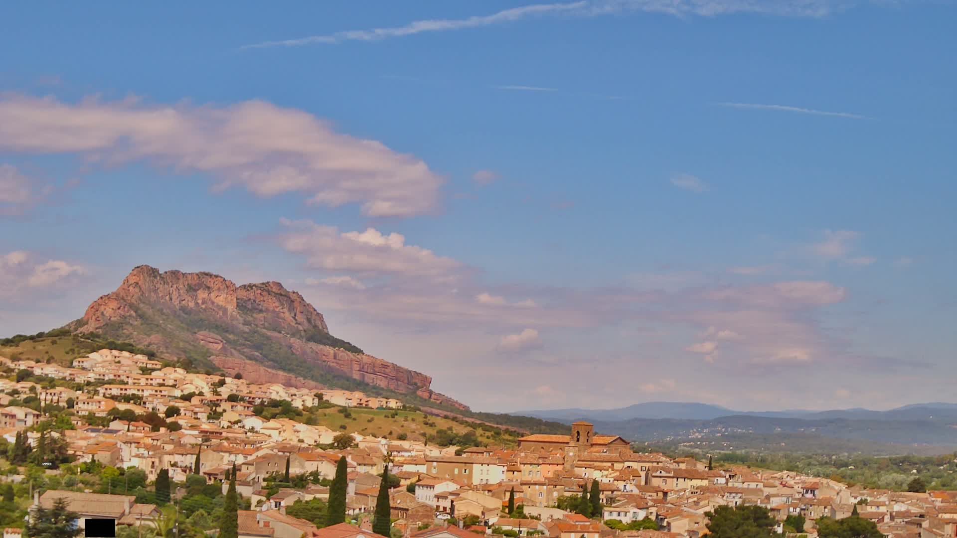 A hilltop town of tan buildings nestles against a reddish-brown rocky mountain under a mostly clear, light-blue sky with scattered clouds.