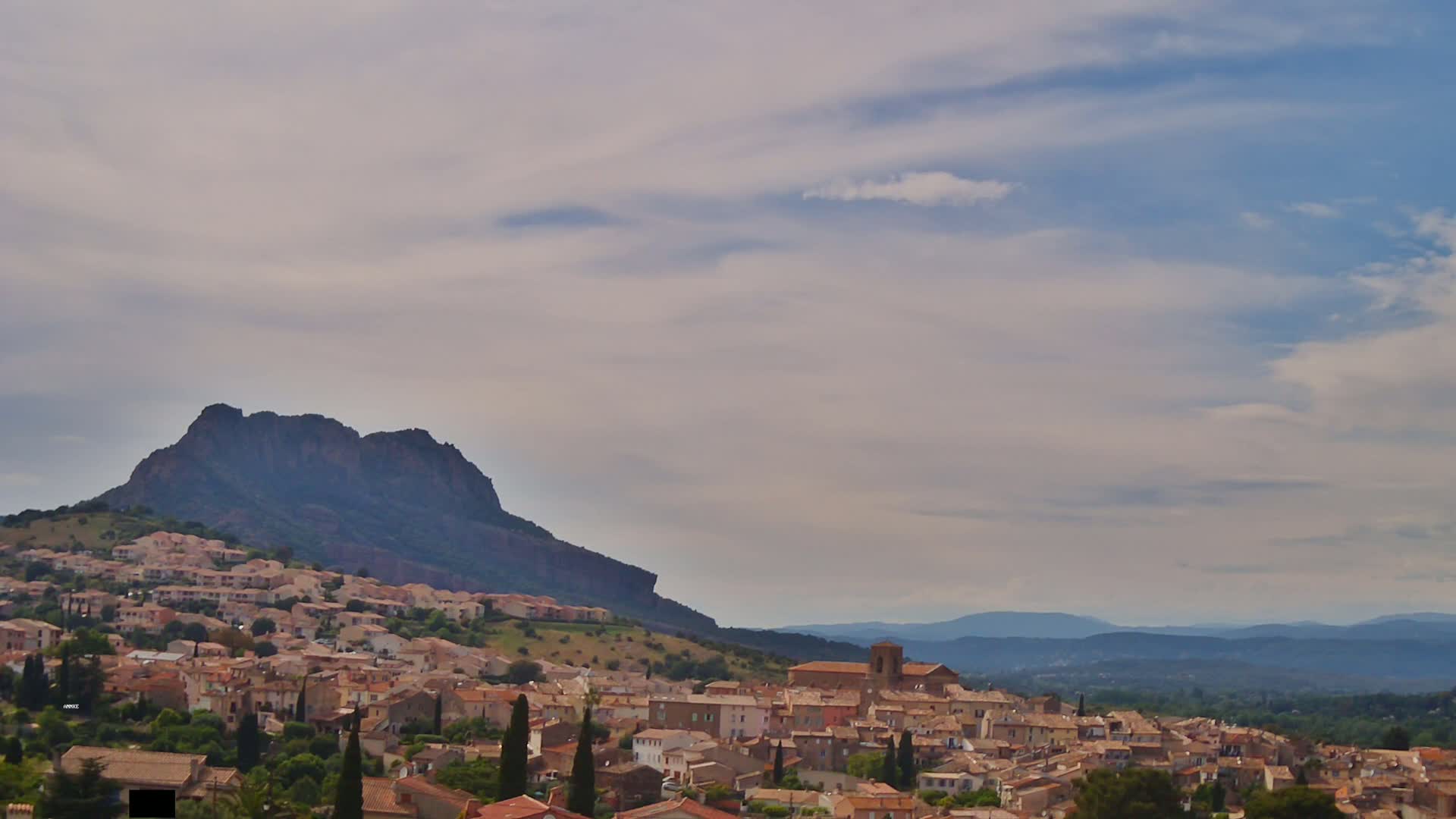 A hilly landscape under a partly cloudy sky shows a village nestled at the foot of a large, dark mountain, with more mountains visible in the distance.